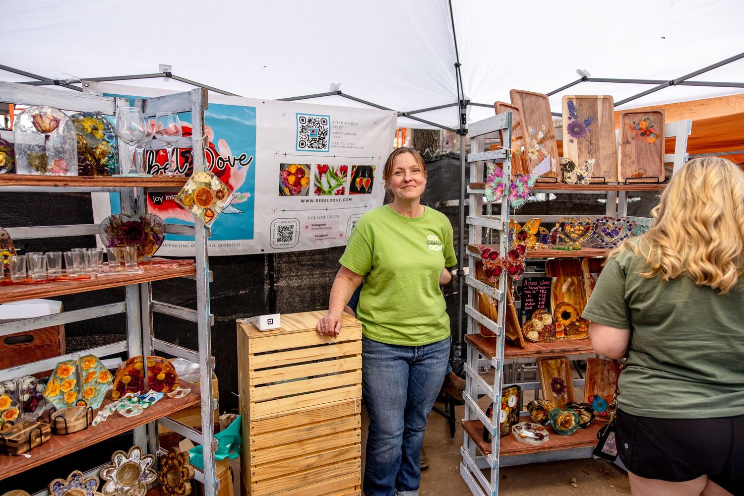 Vendor stands proudly at a colorful craft booth displaying glassware and art at an outdoor market.
