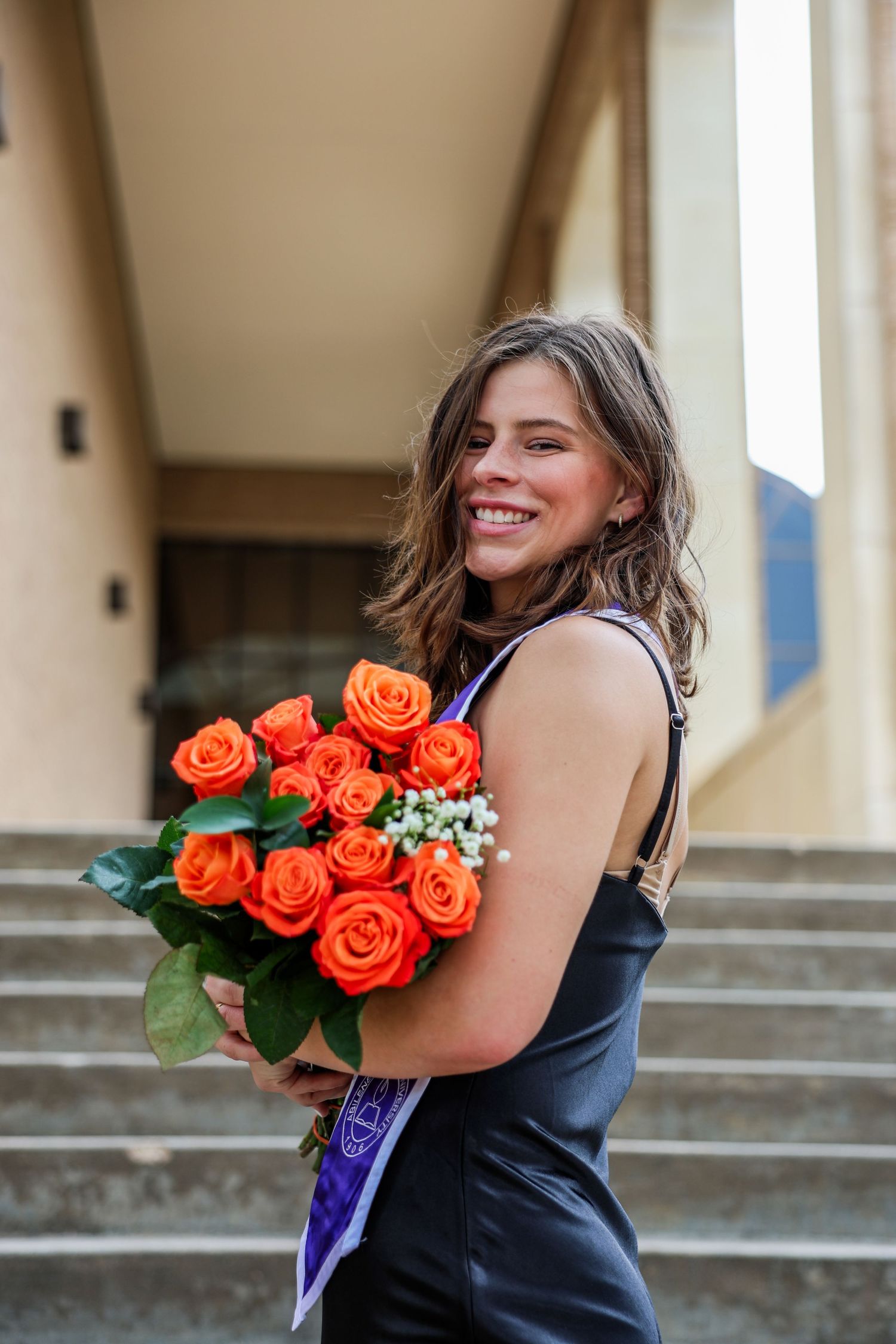 Smiling young woman in black dress holding bouquet of orange roses on outdoor steps.