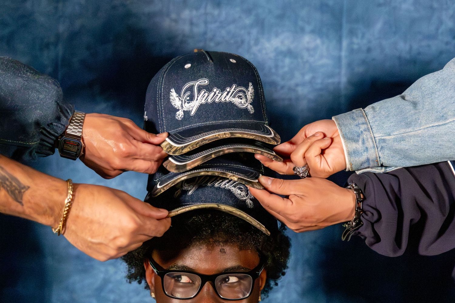 Multiple hands holding stacked Spirit caps above young man with glasses in blue studio.