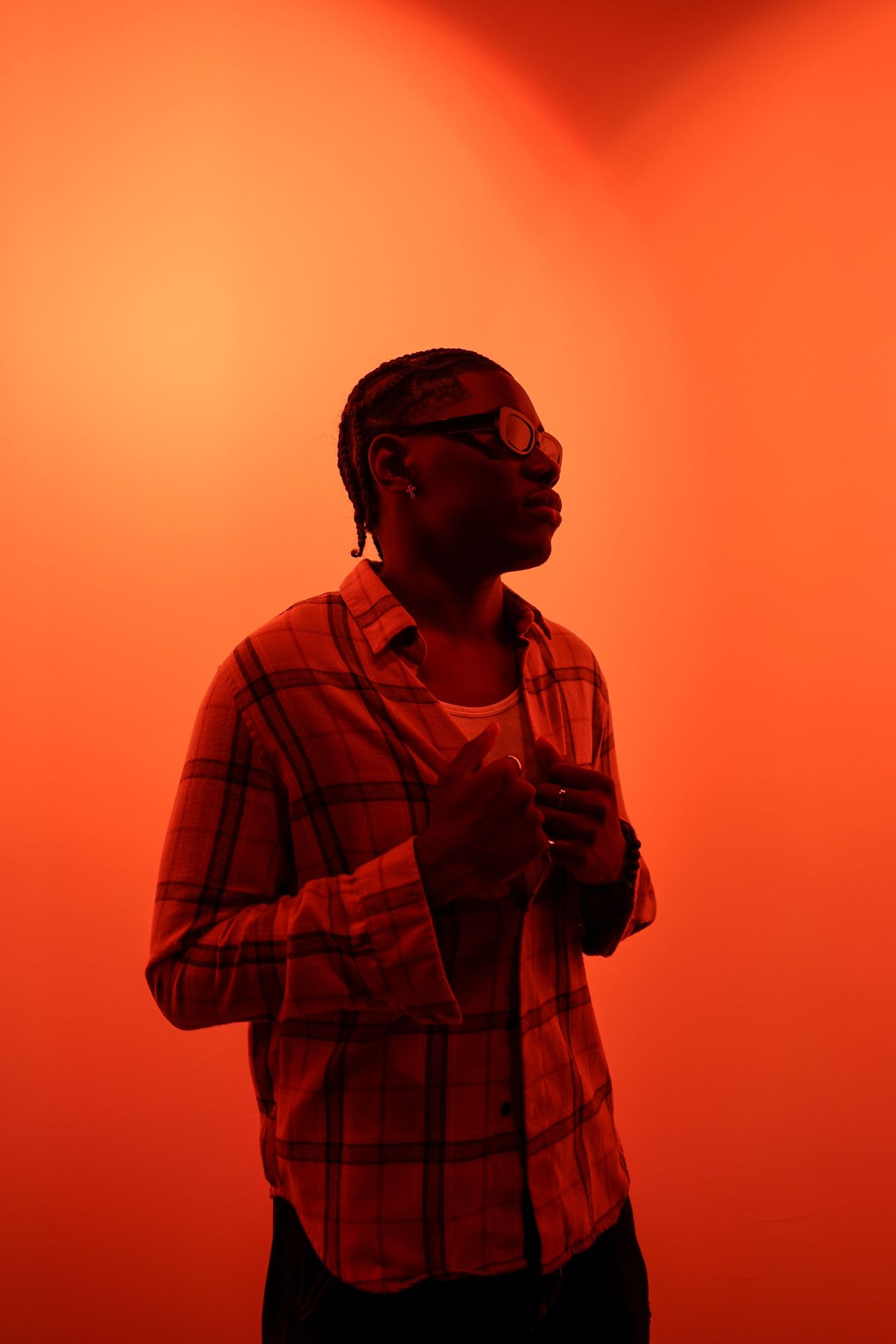 Person in plaid shirt and sunglasses poses in a corner bathed in vivid orange-red light.