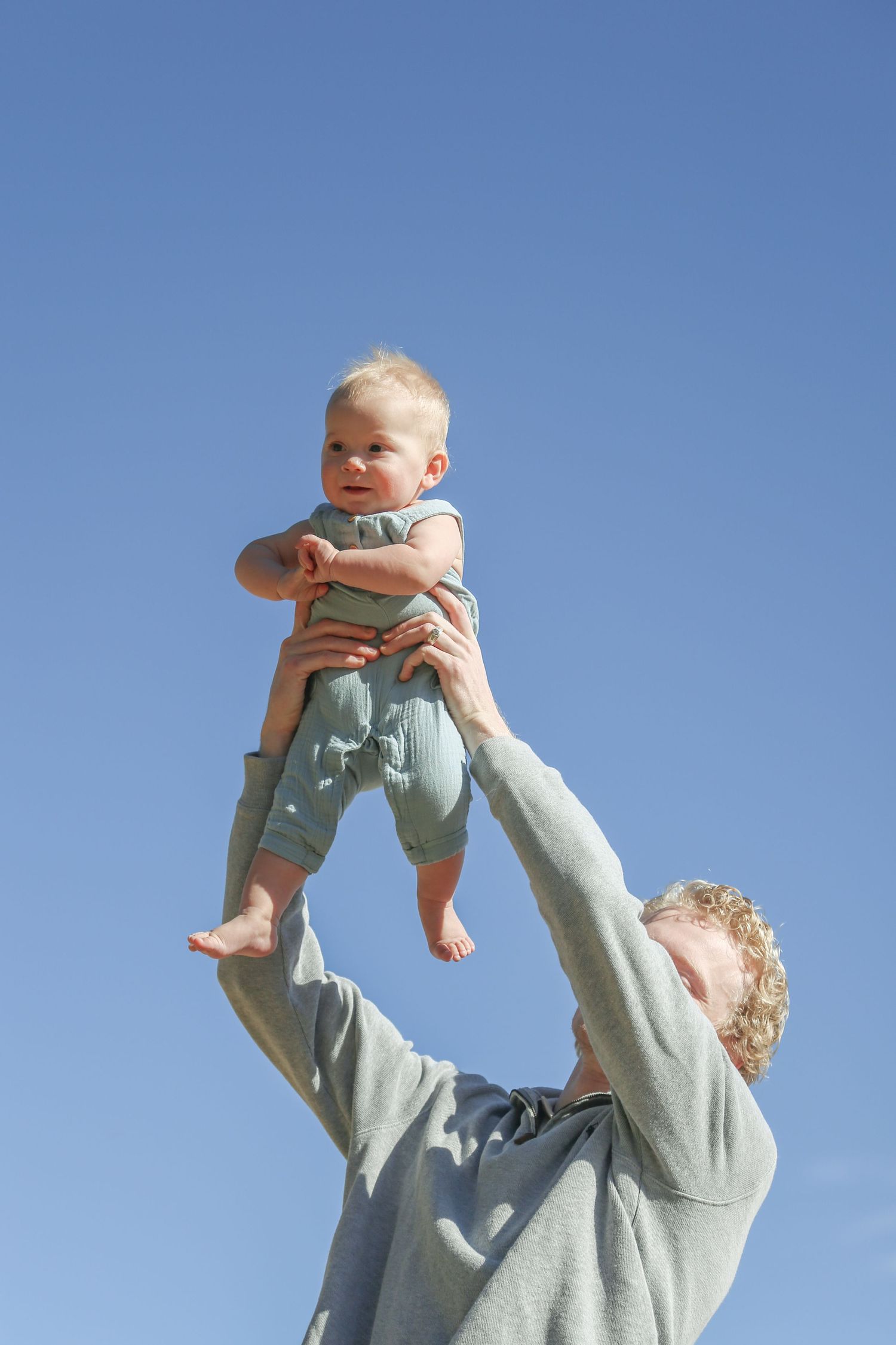 Adult lifting a happy baby in a green onesie high into a clear blue sky outdoors.