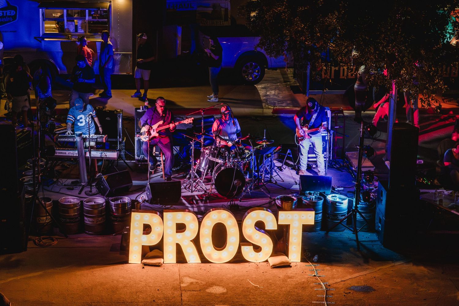Live band performing on stage behind large illuminated PROST letters at an outdoor Oktoberfest event.
