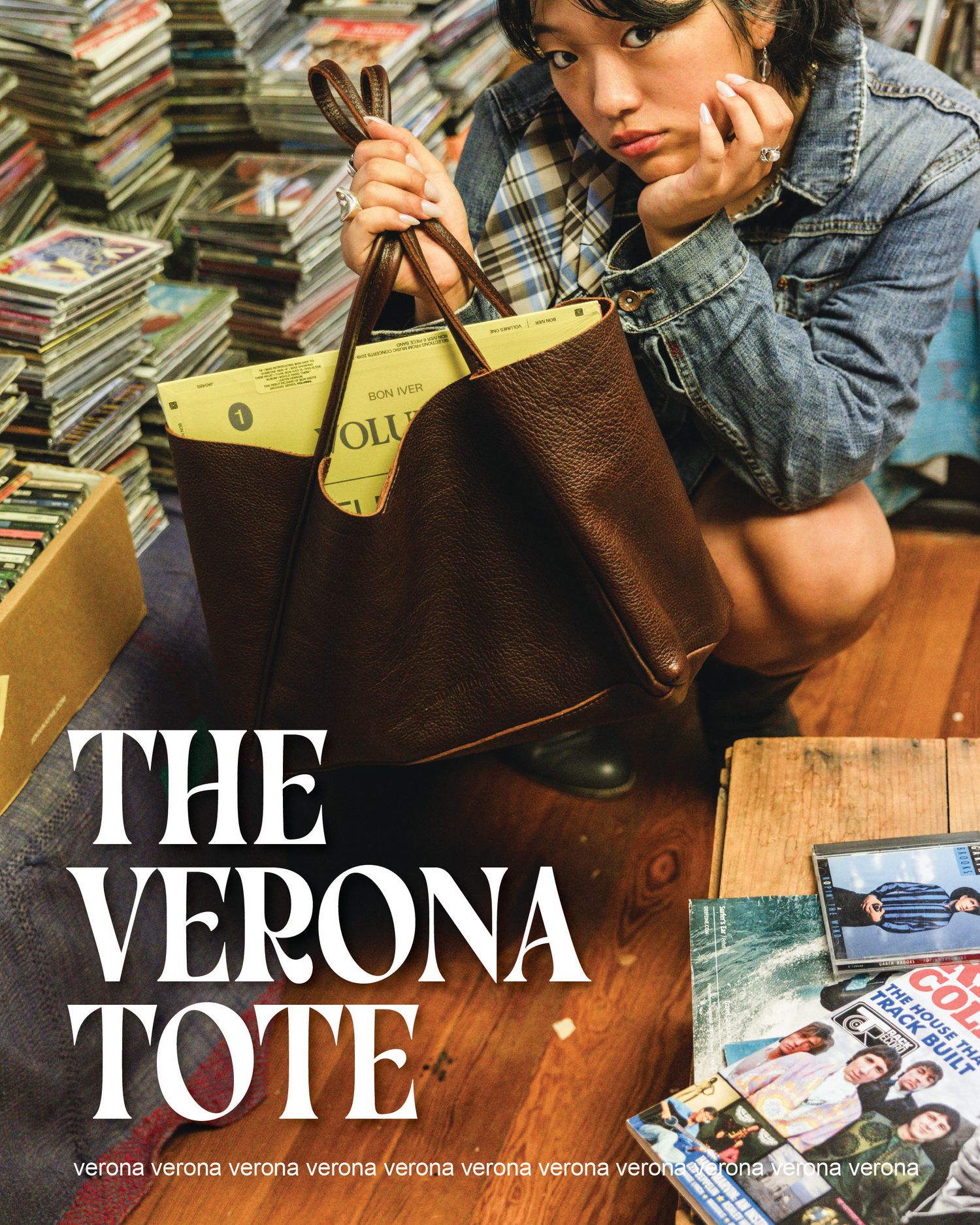 Woman holding the Verona Tote bag, a brown leather tote with tan interior, surrounded by books and magazines.