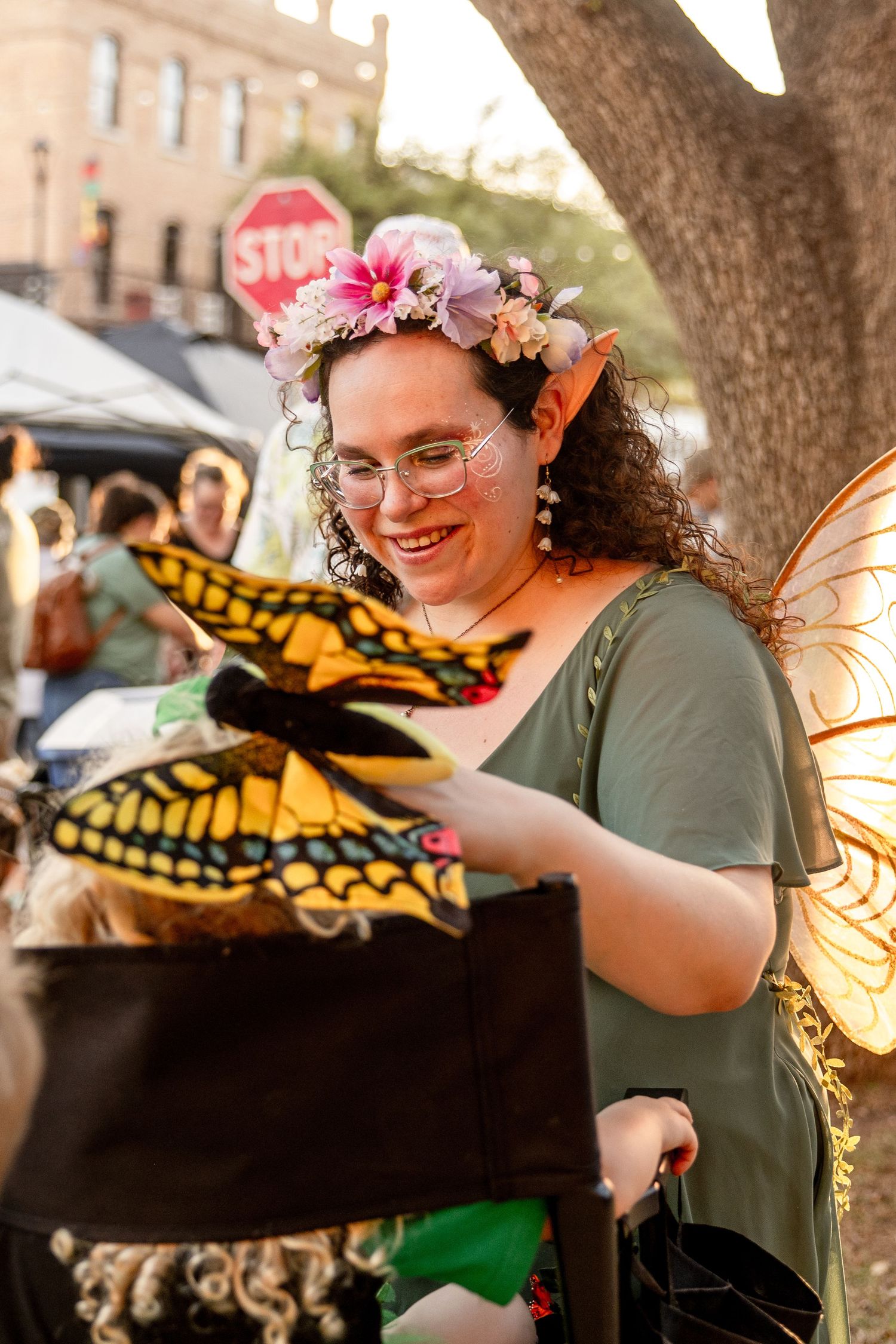 Woman dressed as a fairy with wings and flower crown holds a butterfly at an outdoor event.
