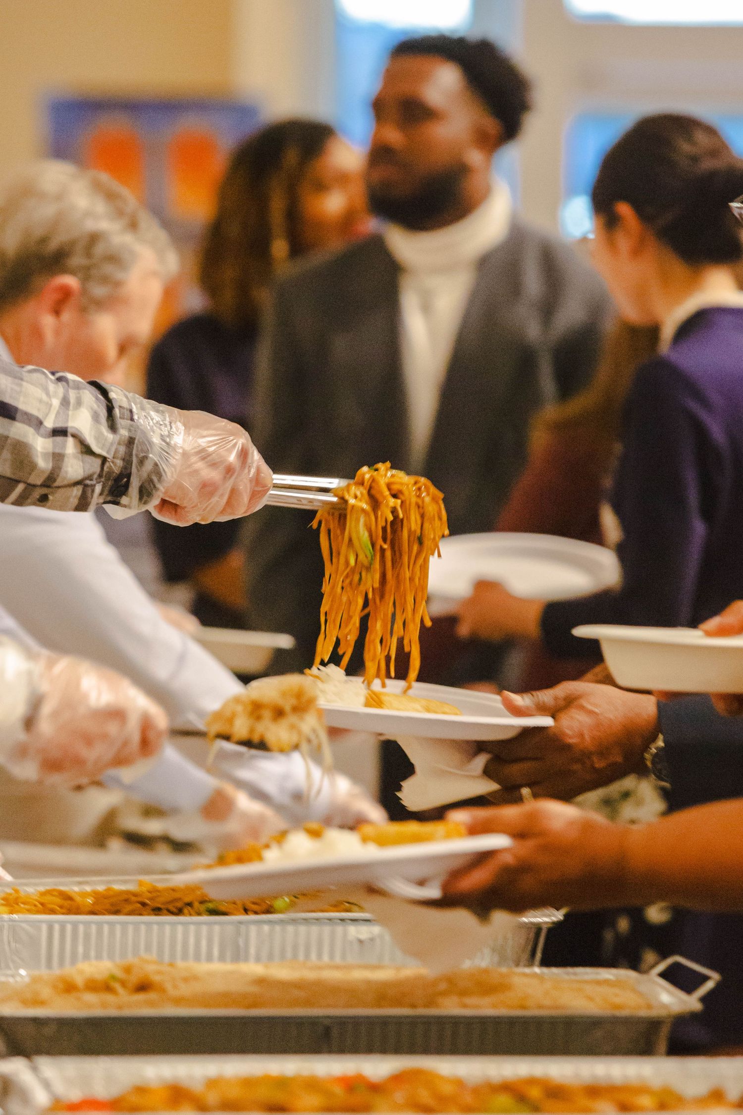 Volunteers serve noodles and food at a buffet line during a multicultural community dining event.