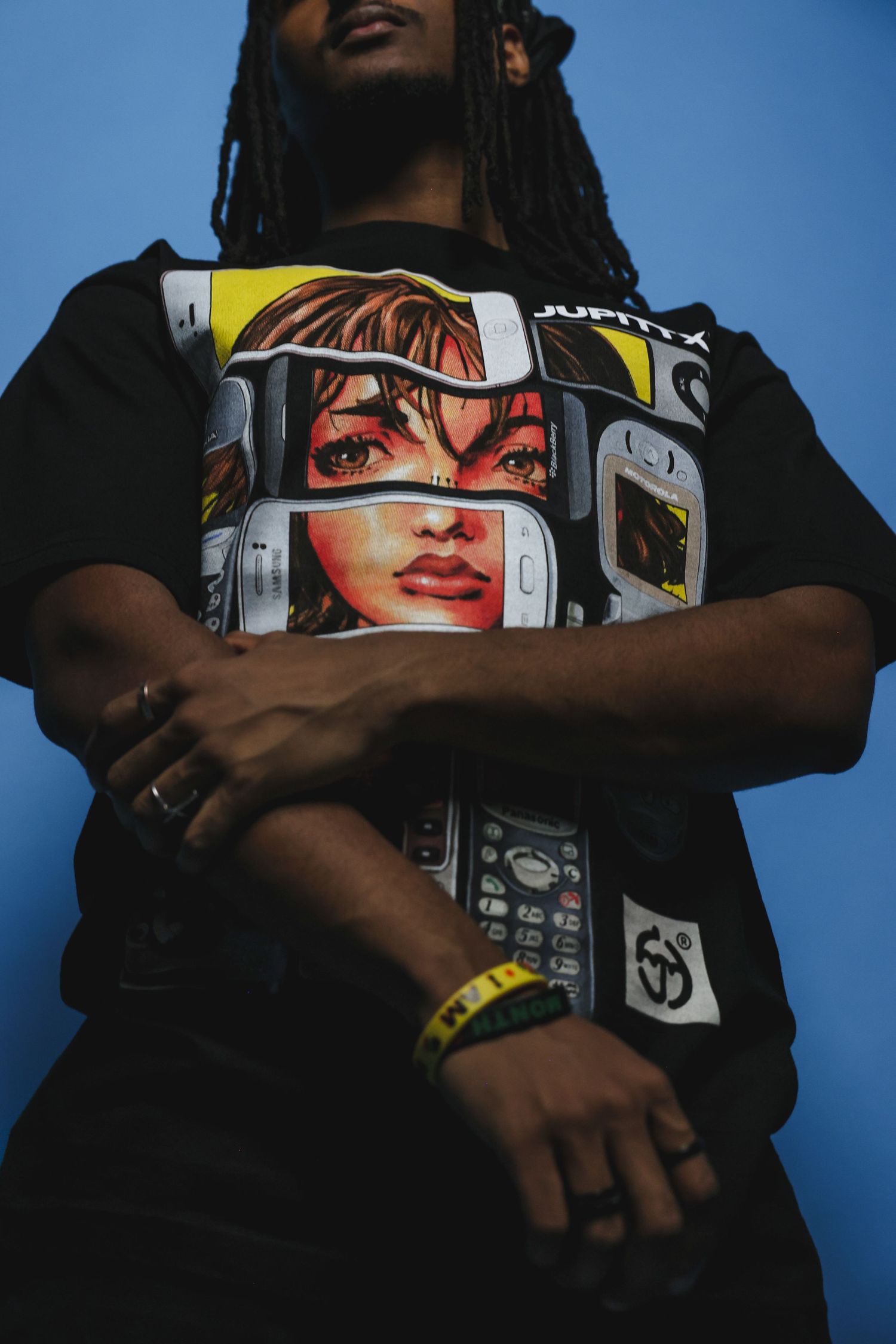 Man with locs crosses arms wearing a bold Jupittar graphic tee against a blue background.