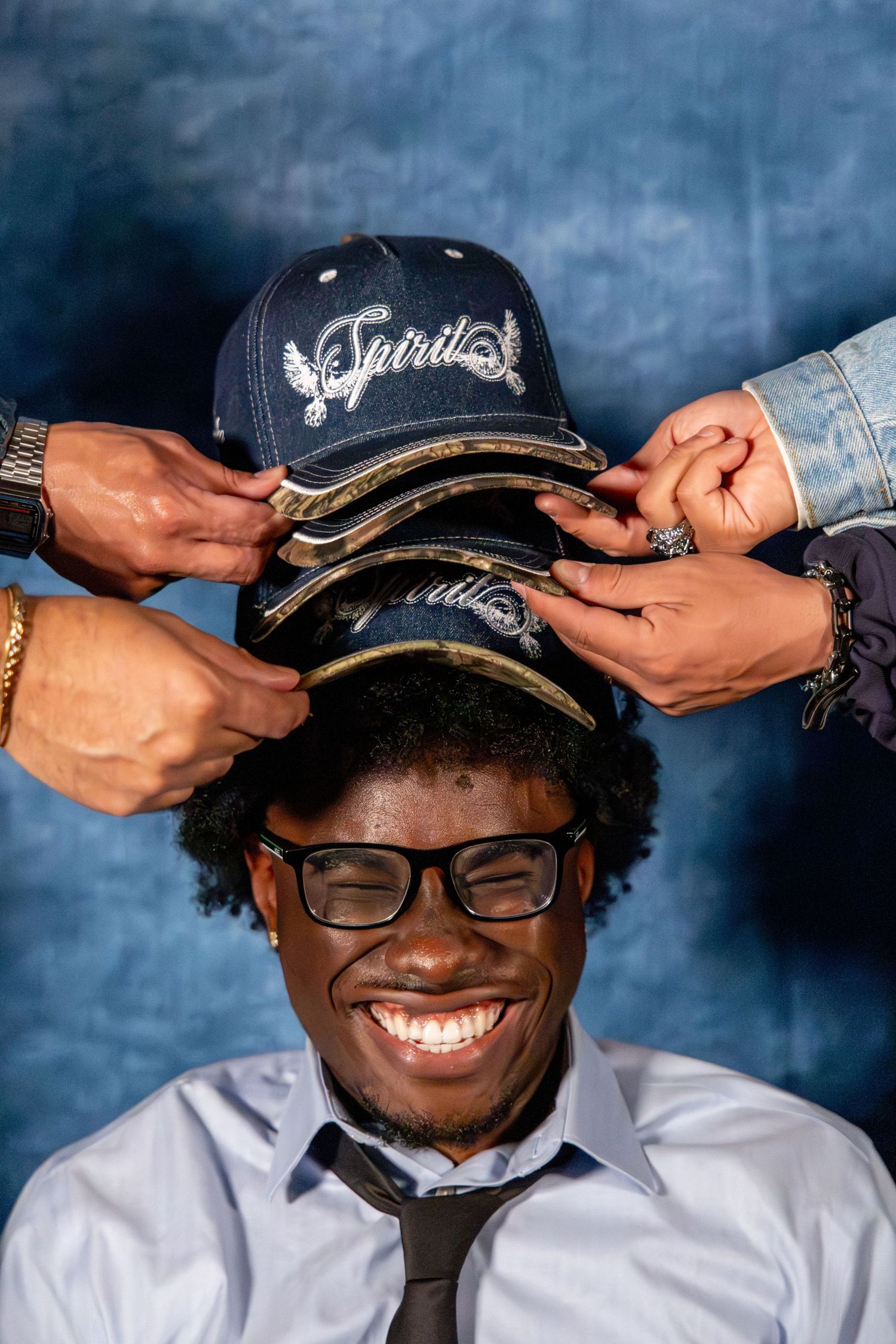 Smiling man in glasses has multiple navy Spirit caps stacked on his head by friends.