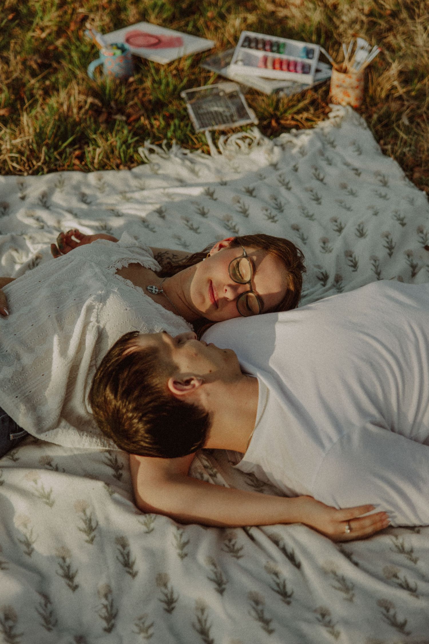 Couple lying on a blanket in a sunny field, relaxing together with art supplies nearby.