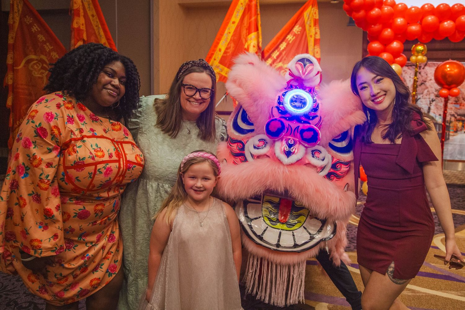 A diverse group of women and a child pose happily with a pink Chinese lion dance costume at a celebration.
