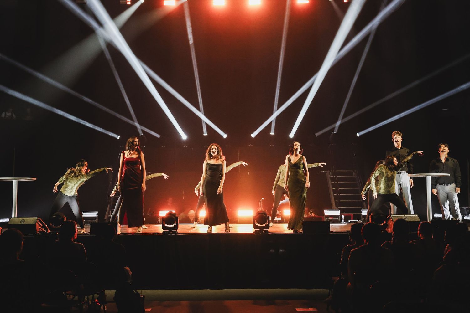 Large ensemble of performers on brightly lit stage with dramatic beam lighting during live show.