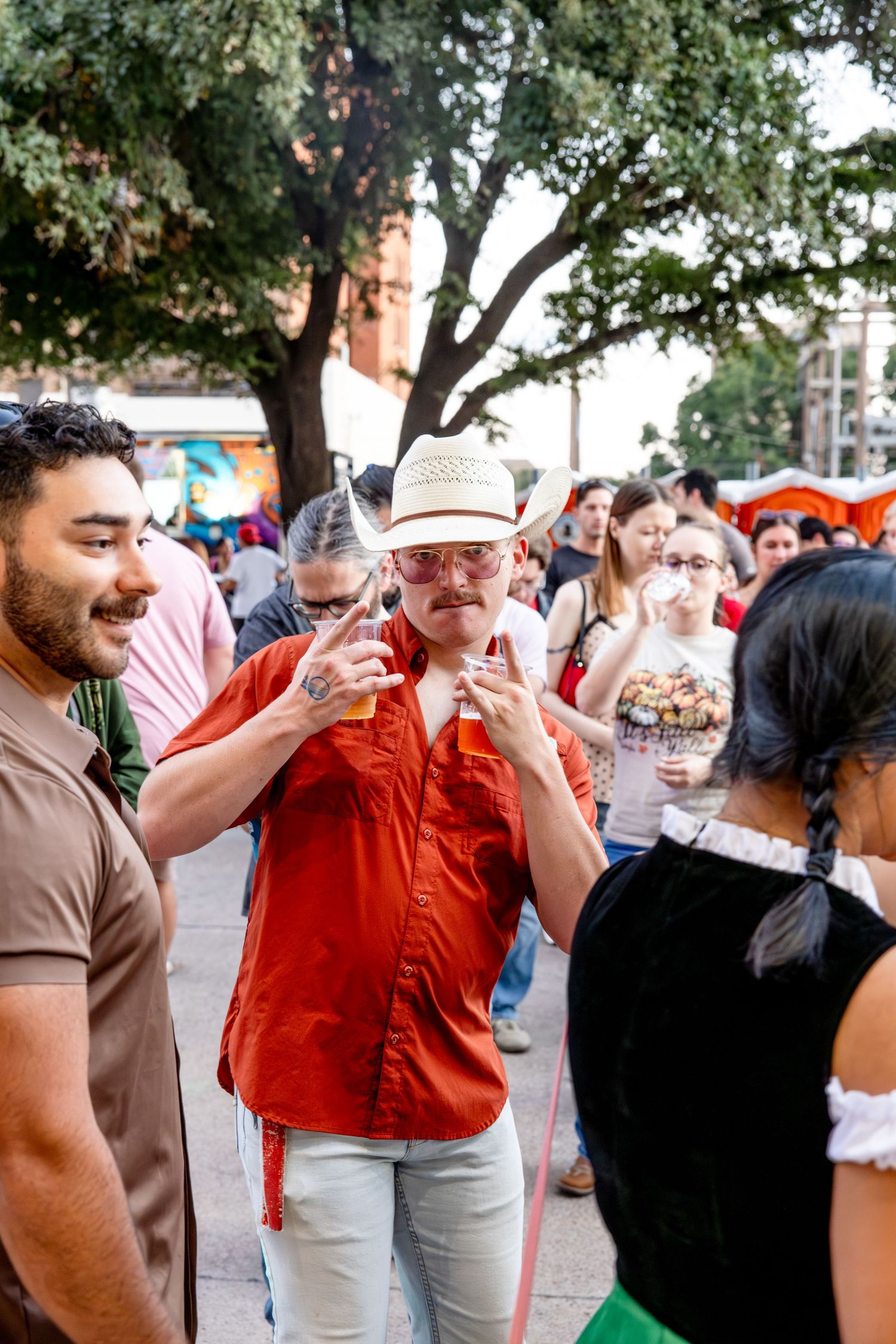 Man in red shirt and white cowboy hat enjoying drinks at a lively outdoor festival.