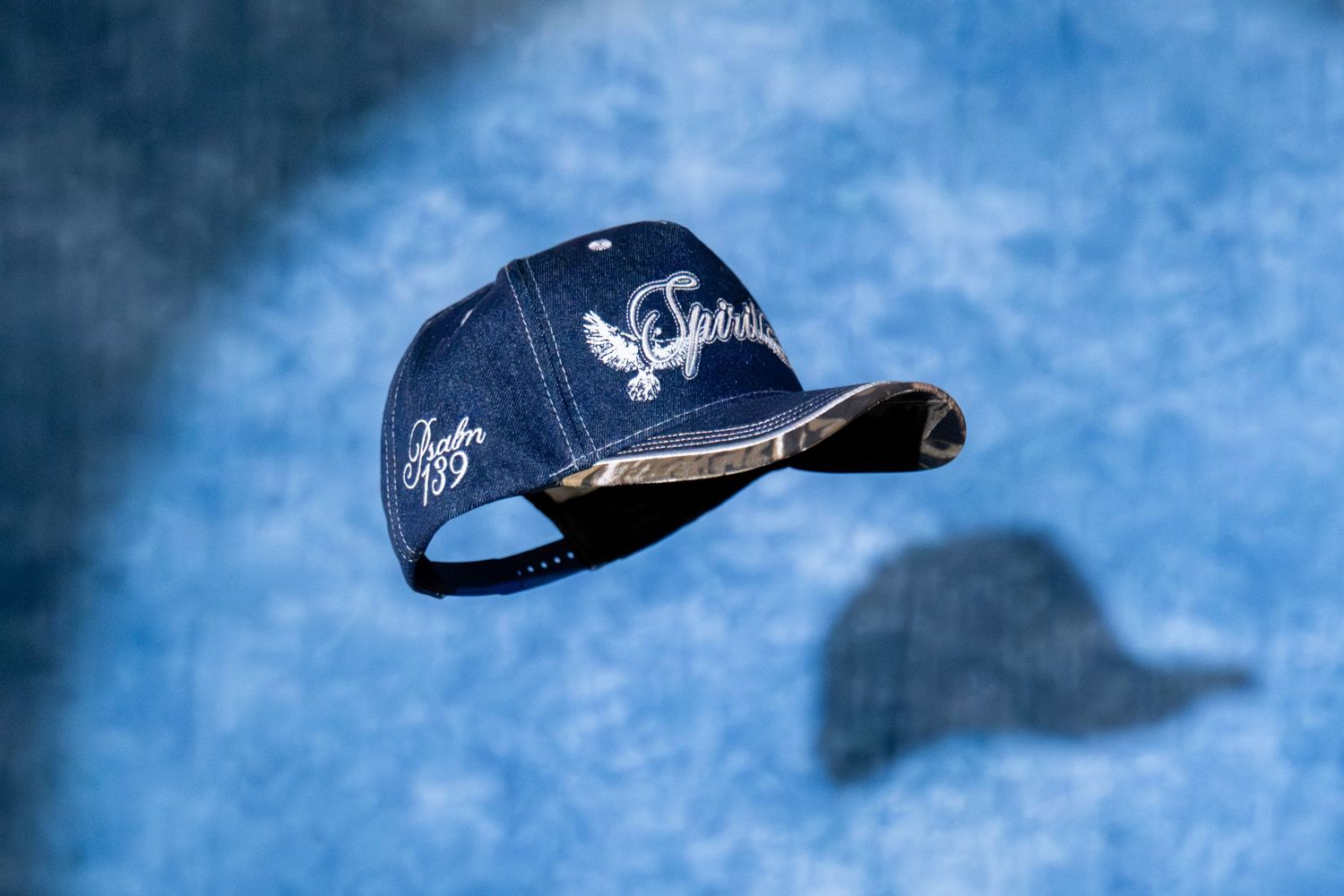 Navy Spirit branded baseball cap floating in mid-air against a blue textured studio background with shadow.