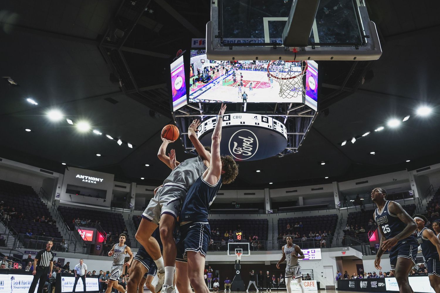Wide-angle view of a college basketball game with players driving to the hoop beneath a large arena scoreboard.