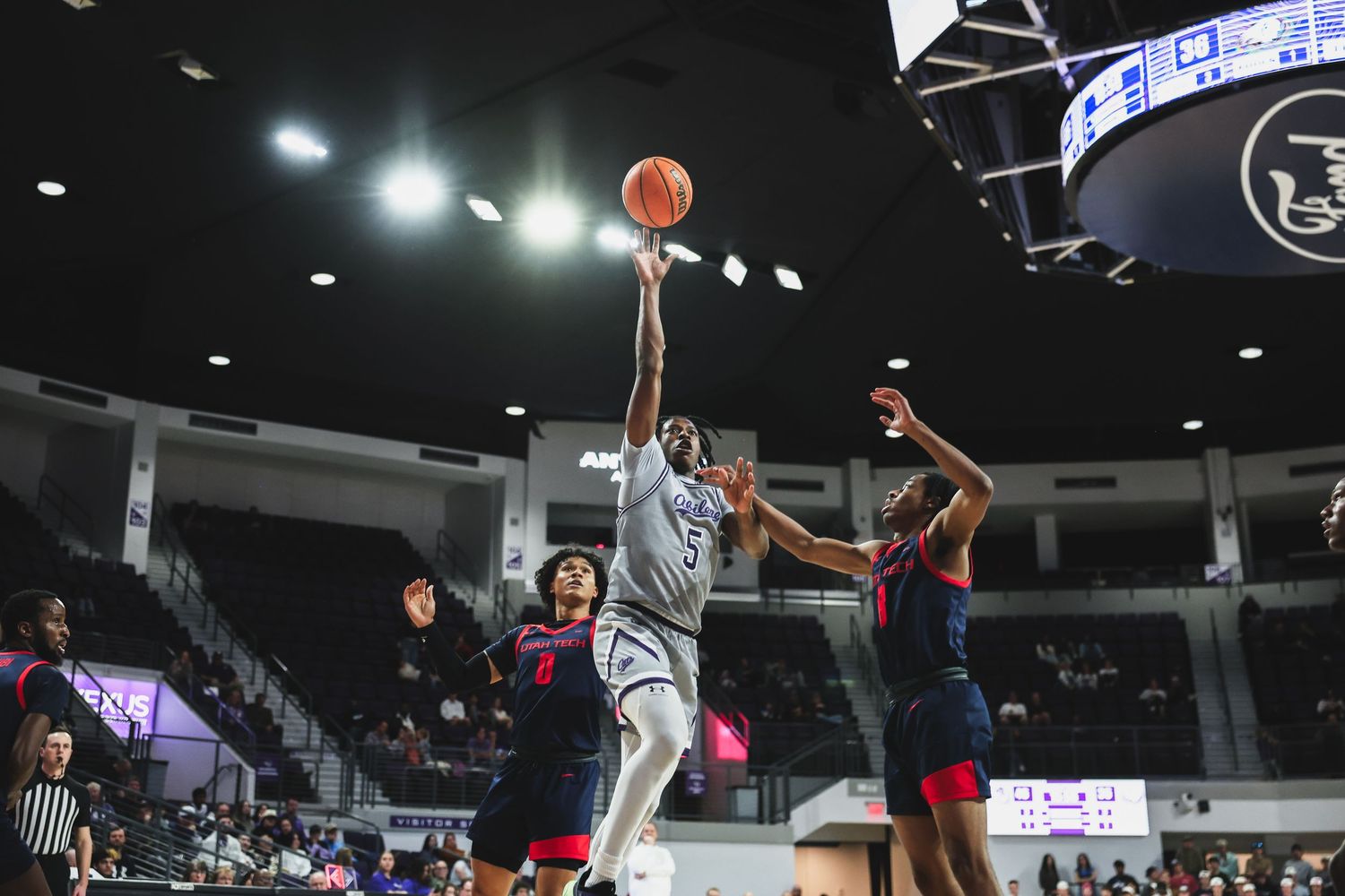 Basketball player in gray uniform shoots a floater between two defenders during a college basketball game.