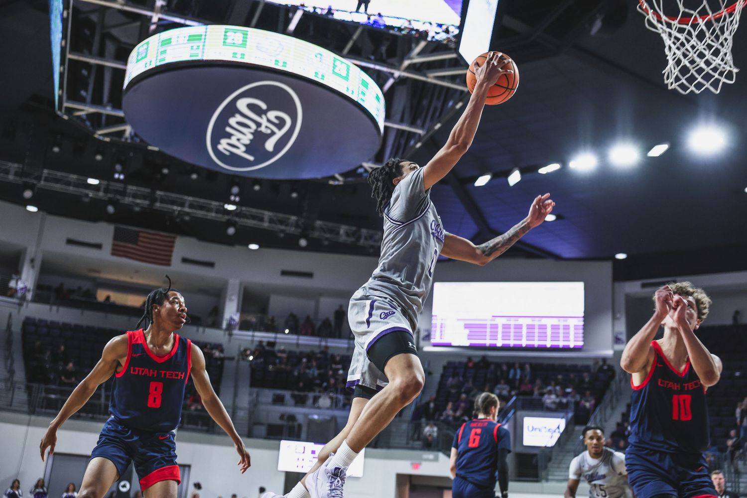 College basketball player in gray uniform leaps for a layup over defenders in a well-lit arena.