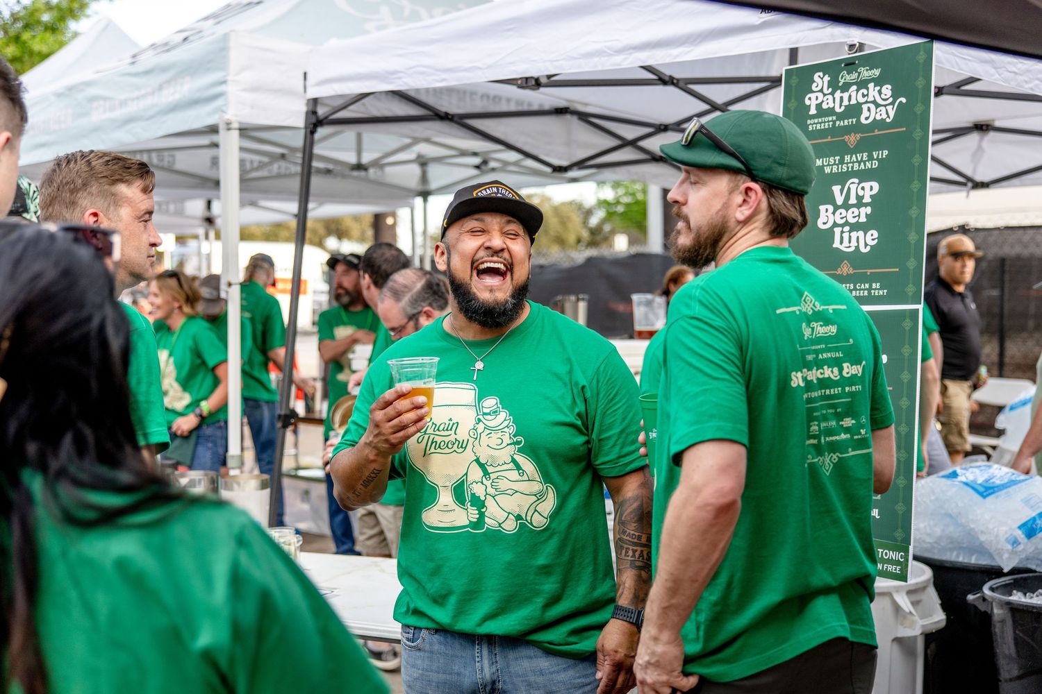 Men in green St. Patrick's Day shirts laughing and enjoying drinks at an outdoor beer festival.