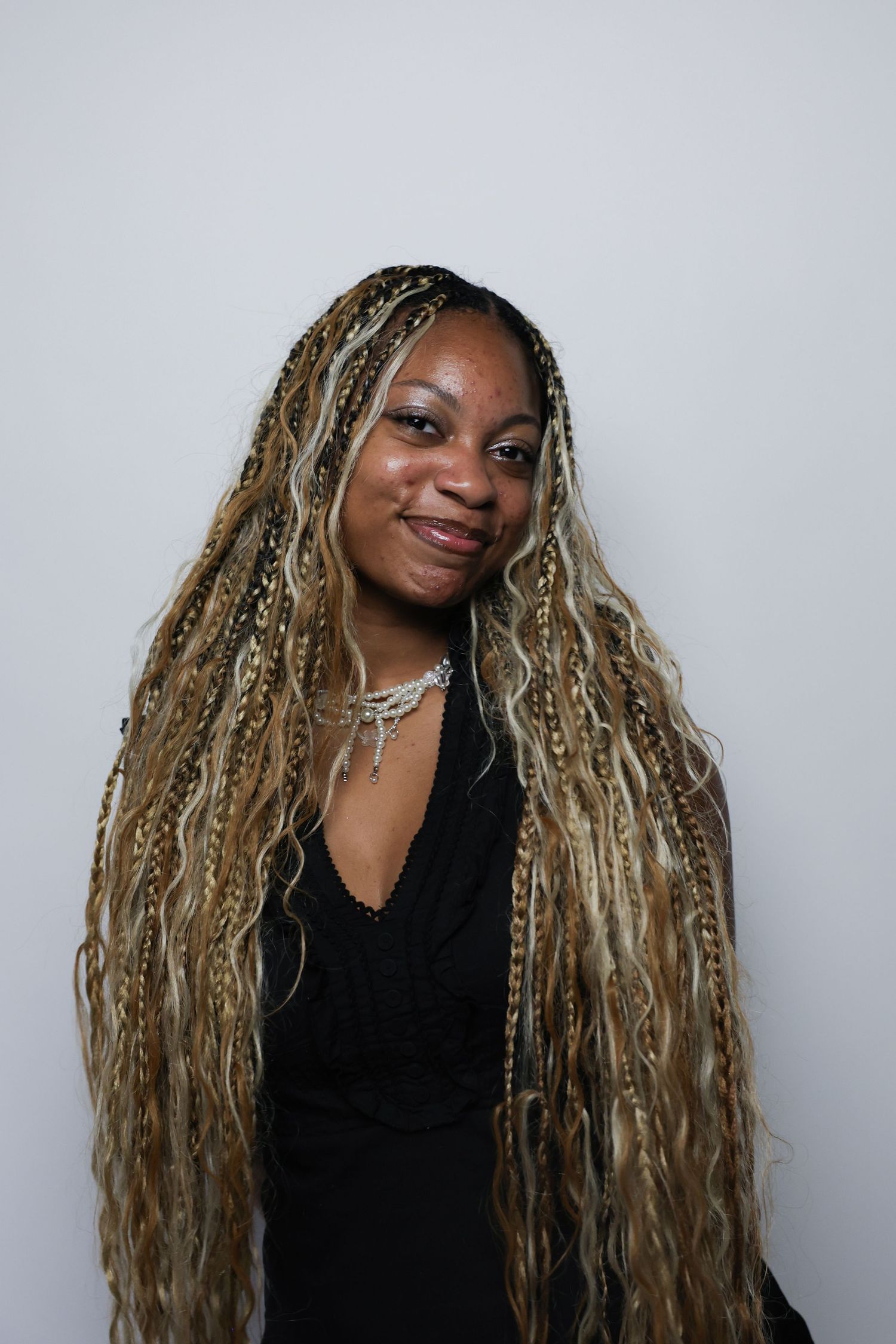 Smiling woman with long blonde braids wearing black top and layered necklaces on white background.
