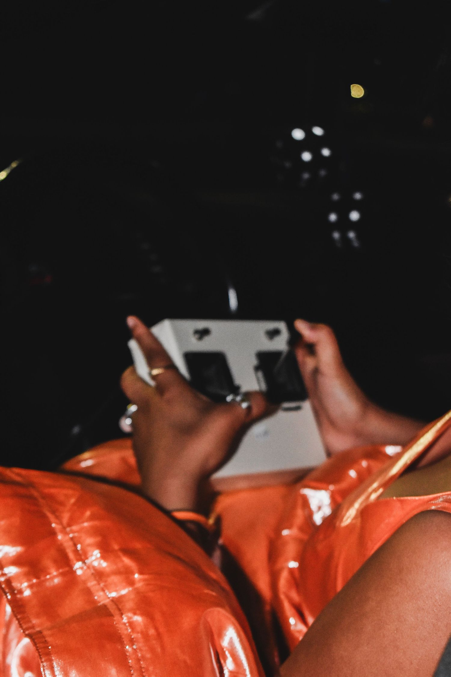 A person in an orange shiny outfit holds a Polaroid camera in their lap at night.