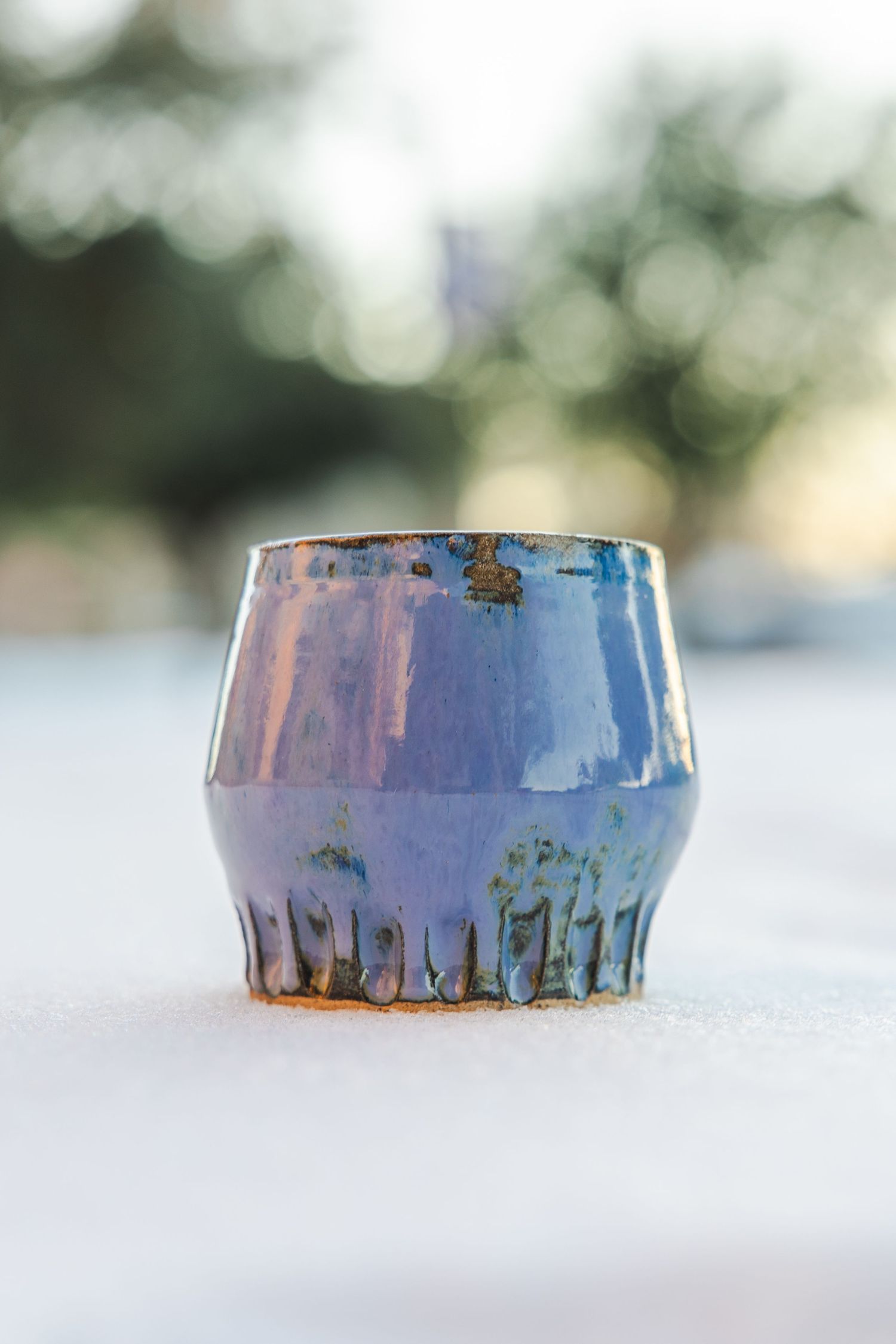 Blue iridescent glazed handmade ceramic tumbler sitting on snow with bokeh background.