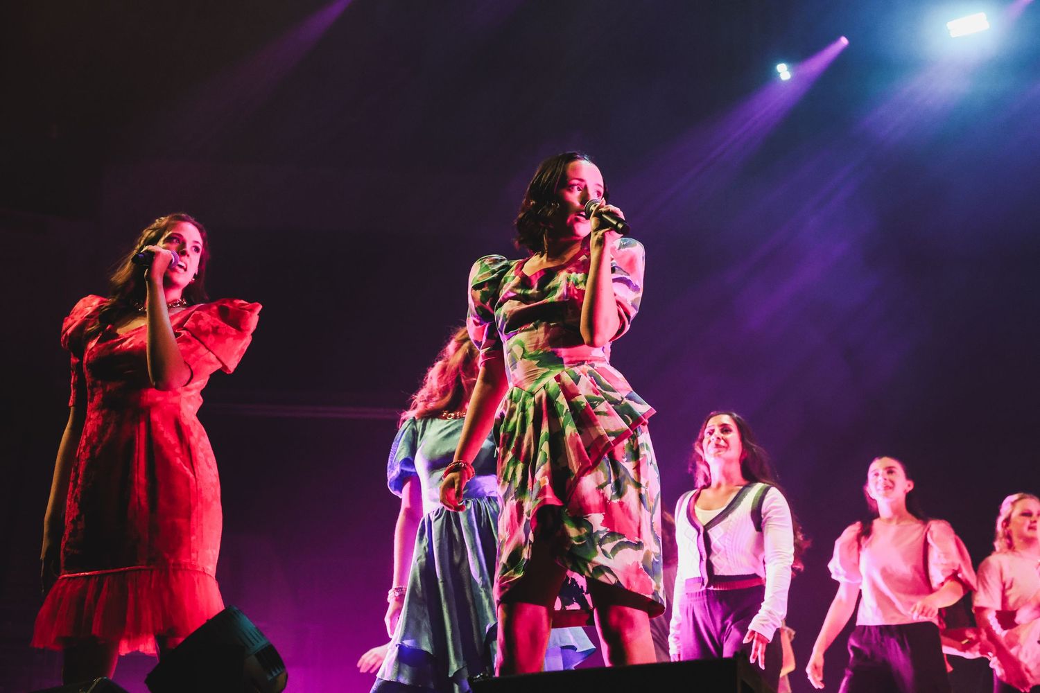 Women performers on stage in colorful outfits under pink and purple concert stage lighting.
