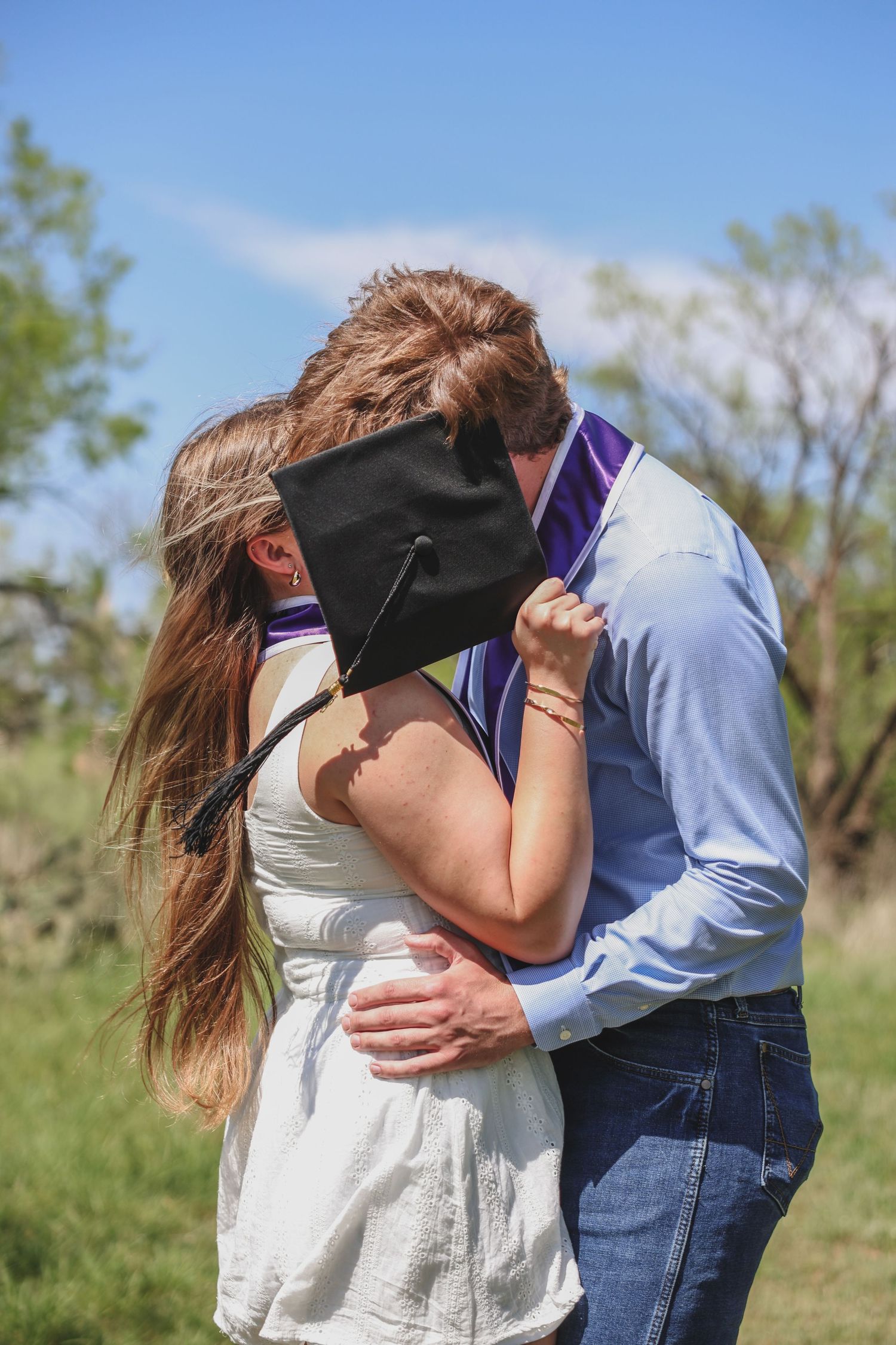 Couple sharing a kiss hidden behind a black graduation cap outdoors in a green field.