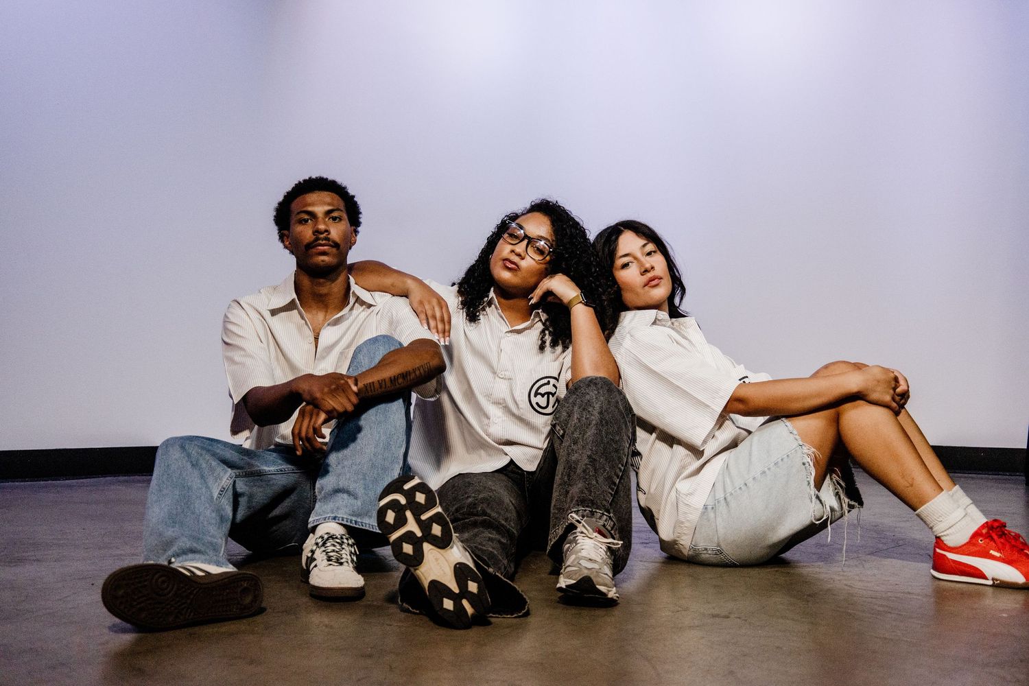 Three friends in casual streetwear sit together on floor against white wall for fashion shoot.