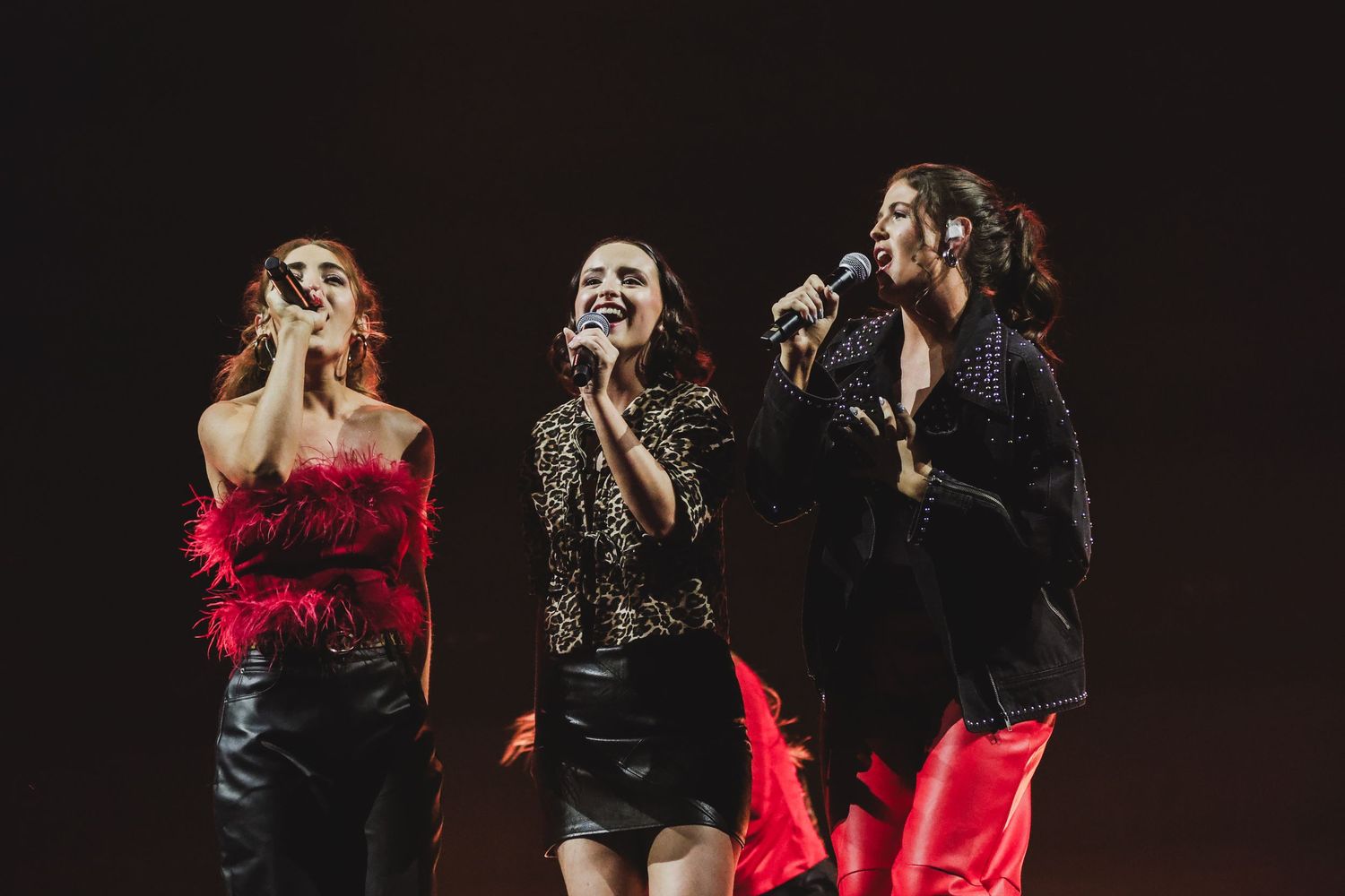 Three women singing on stage with microphones under warm spotlight during live concert performance.