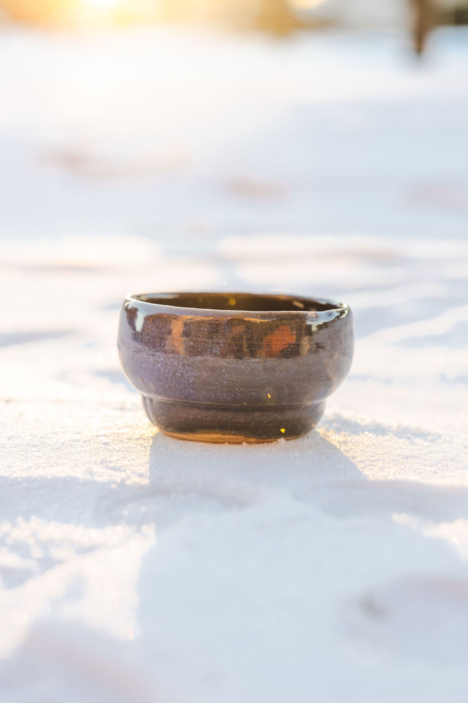 Brown glazed handmade ceramic bowl on snow with warm golden sunset light reflection.