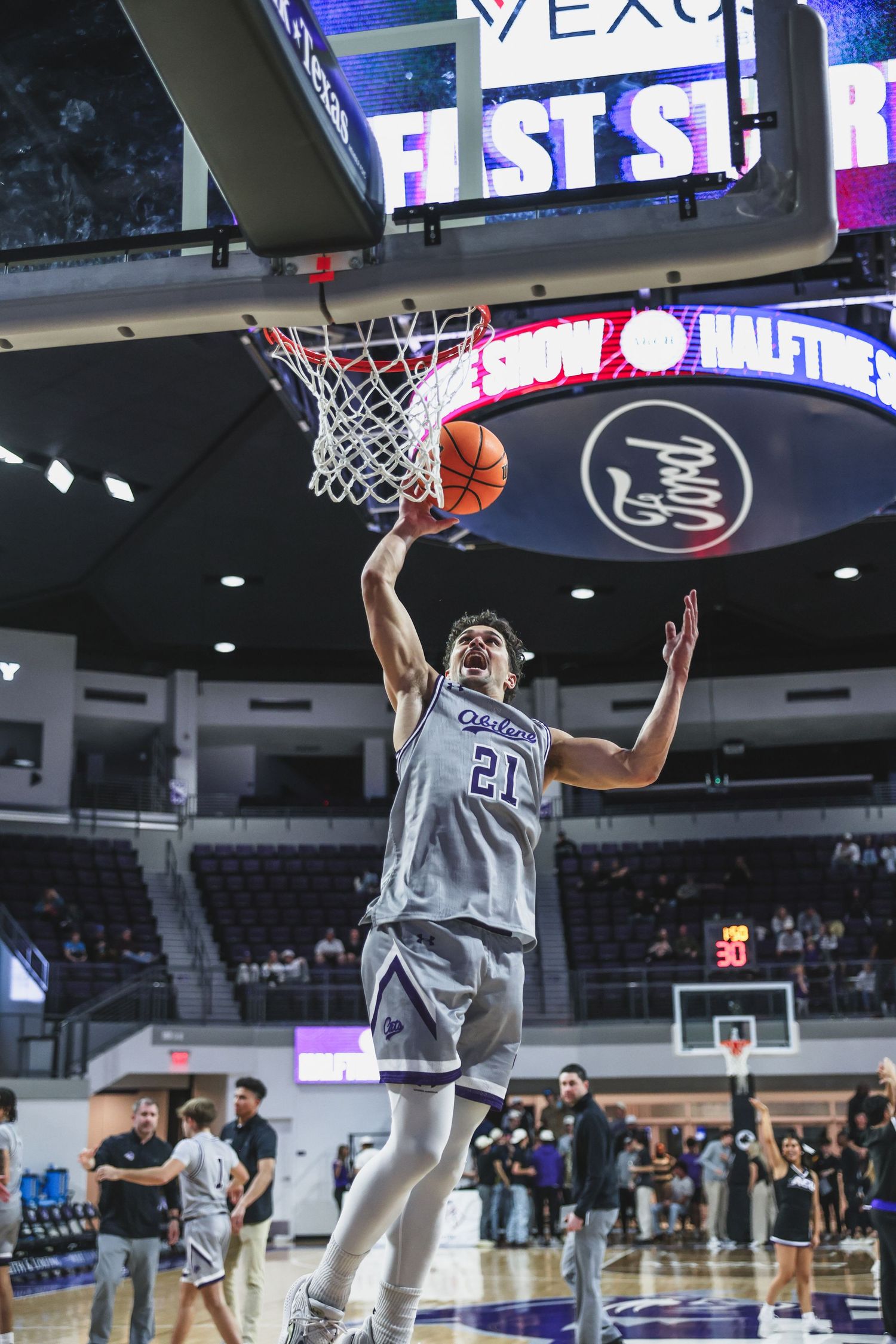 College basketball player #21 in gray uniform practices a layup during halftime warmups in a modern arena.