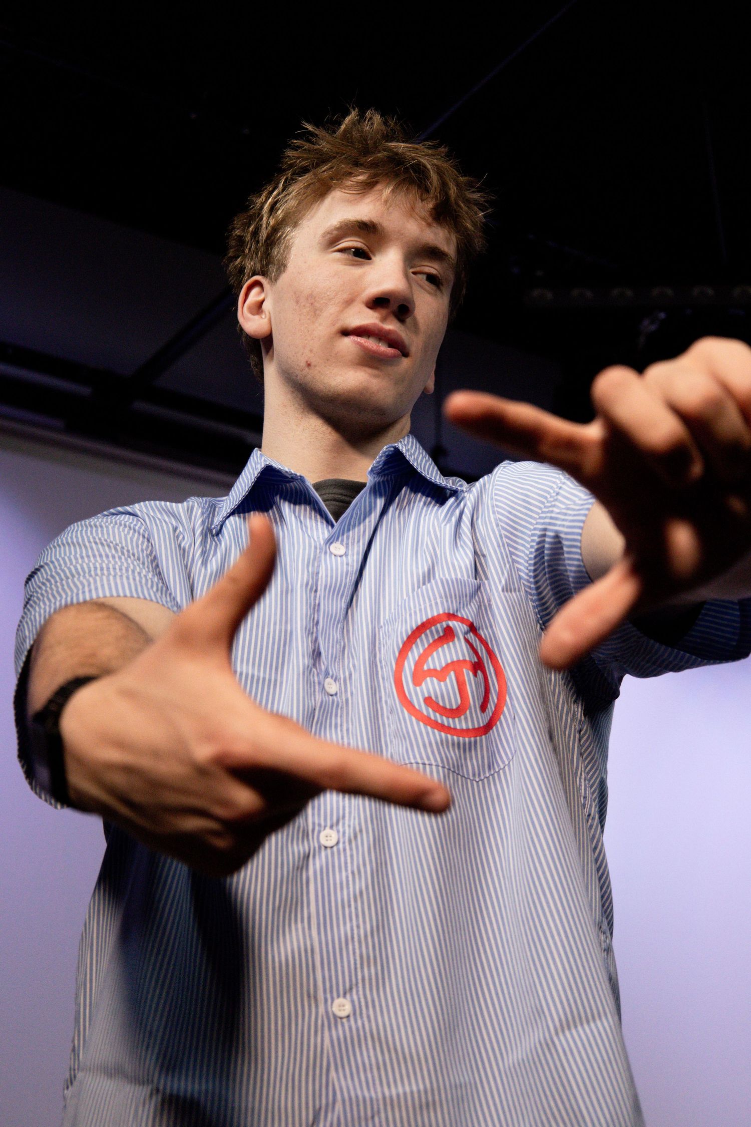 Young man in blue striped shirt with red logo gestures toward camera in dynamic pose.