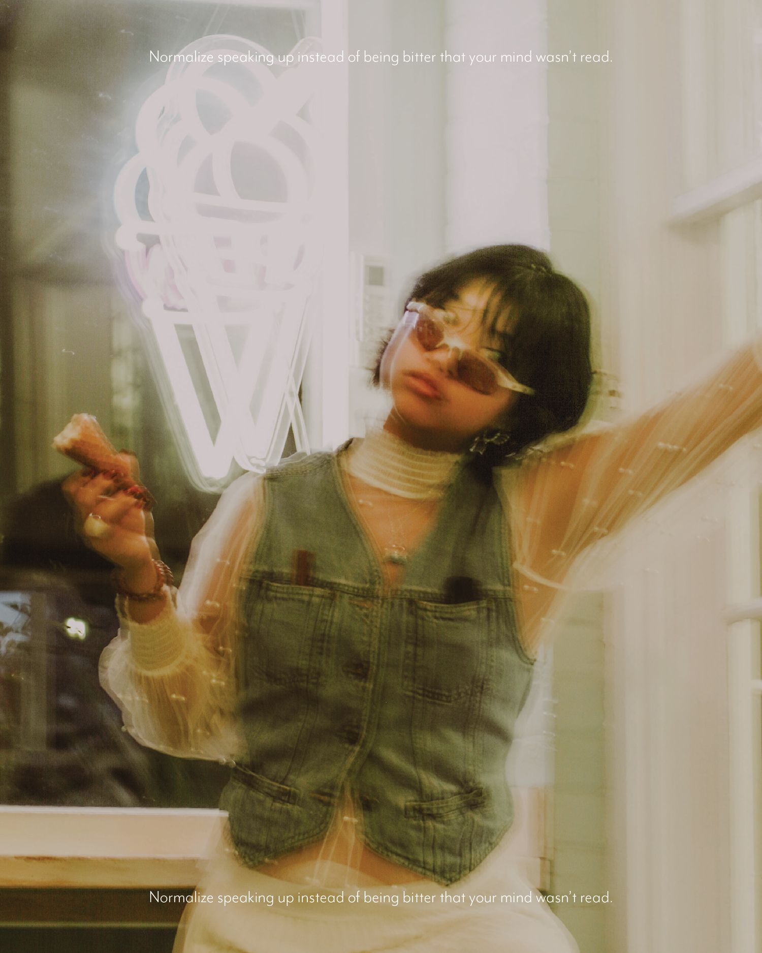 Young woman in denim overalls and tinted glasses poses artistically near a neon sign in a dreamy, film-inspired photo.