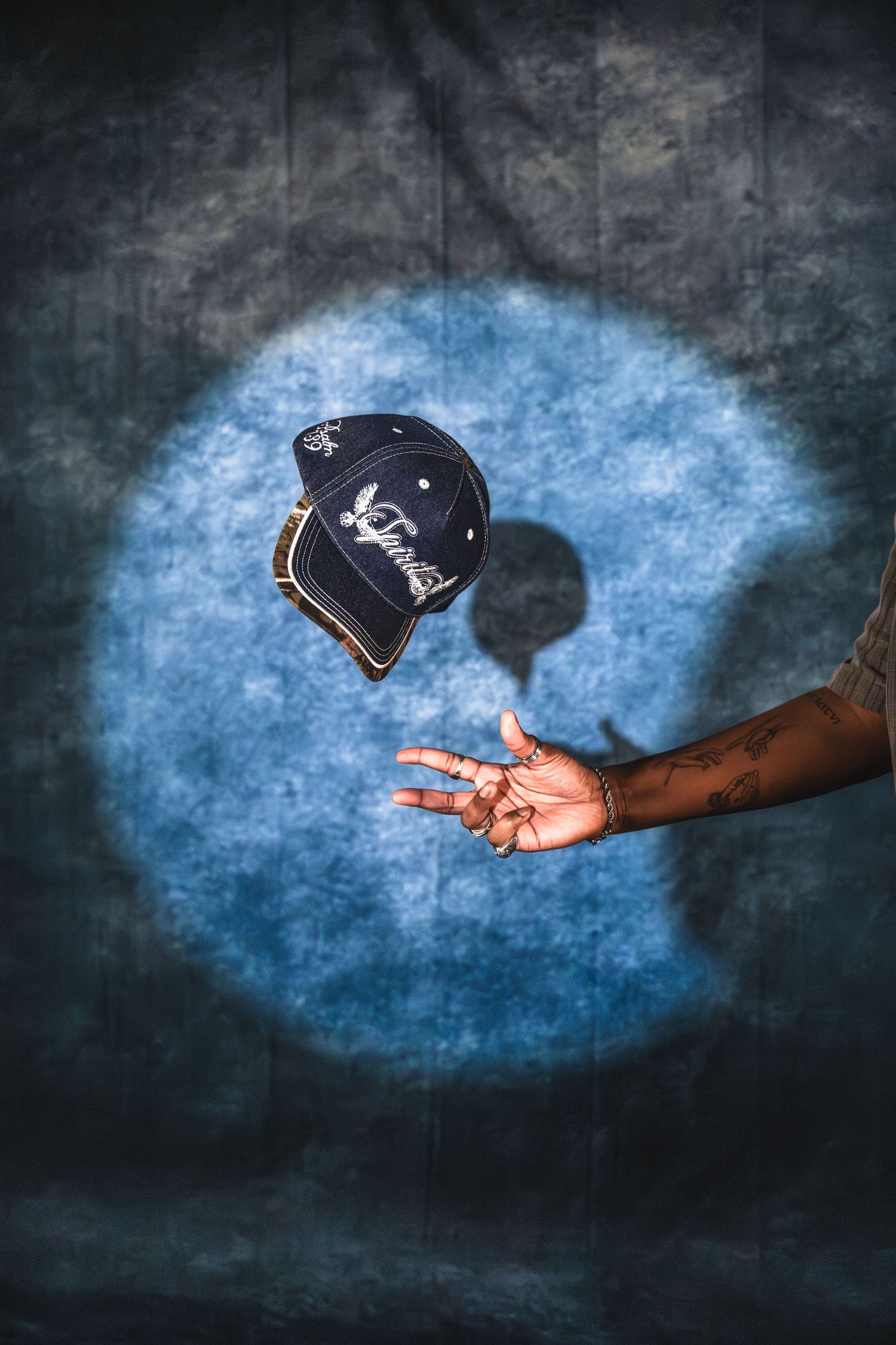 A navy blue cap tossed in the air above a tattooed hand against a blue backdrop.