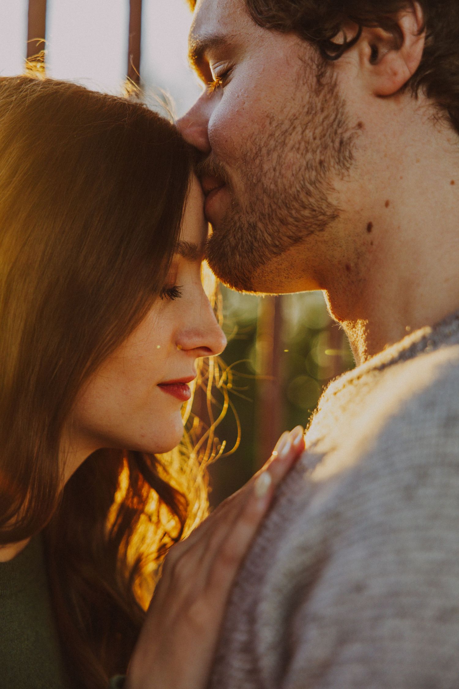 Romantic close-up of couple at golden hour, man kissing womans forehead in warm sunlight.