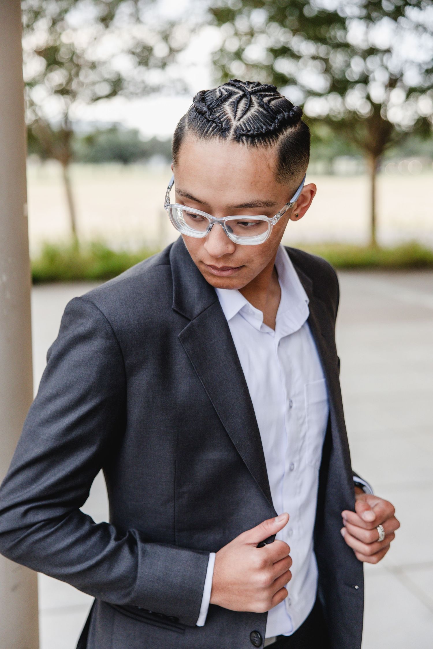 Young man in gray suit with braided hair and clear glasses adjusting jacket outdoors.