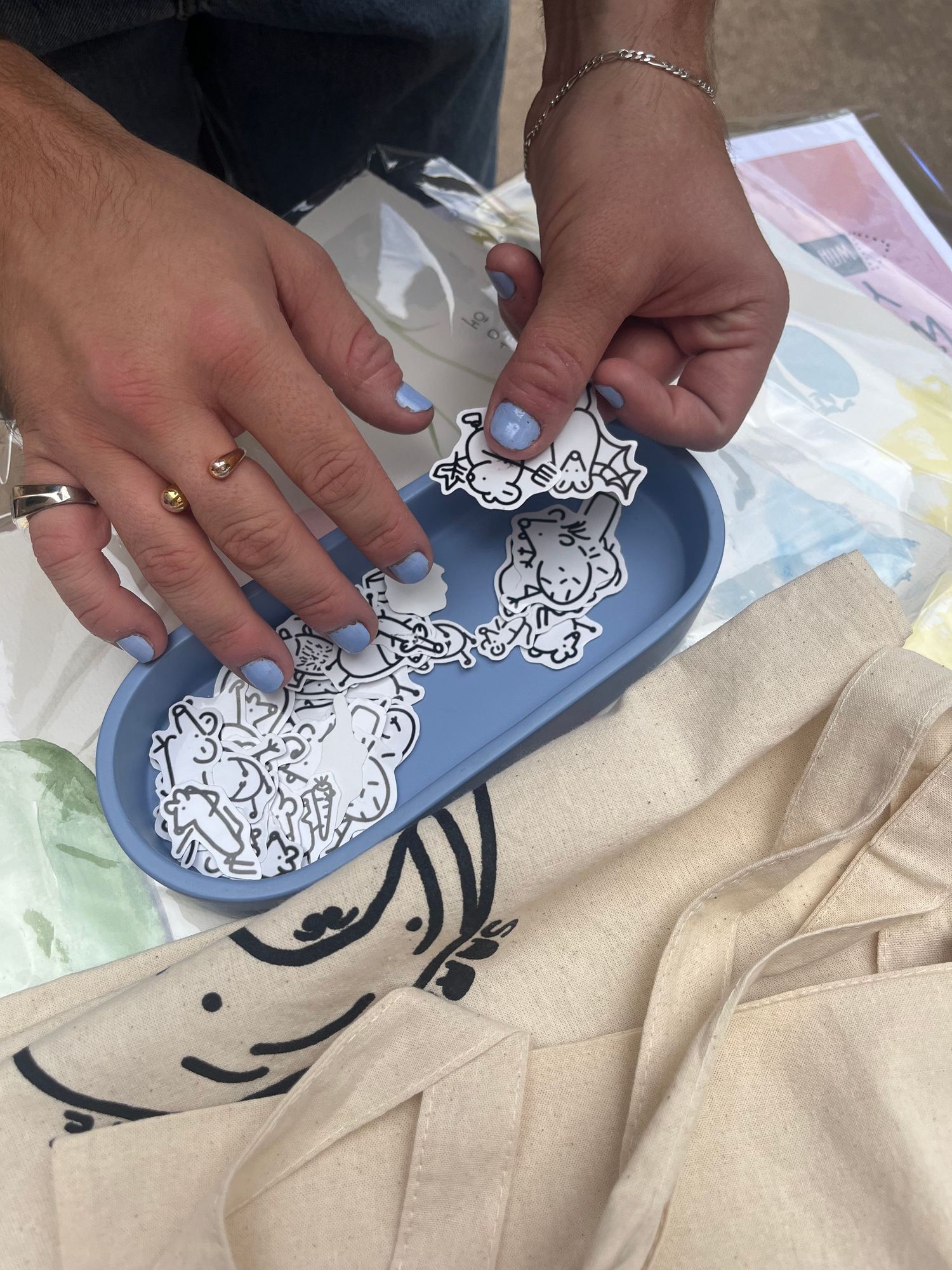 Hands with blue nails sorting small cartoon rat stickers on a blue tray next to printed canvas tote bags.