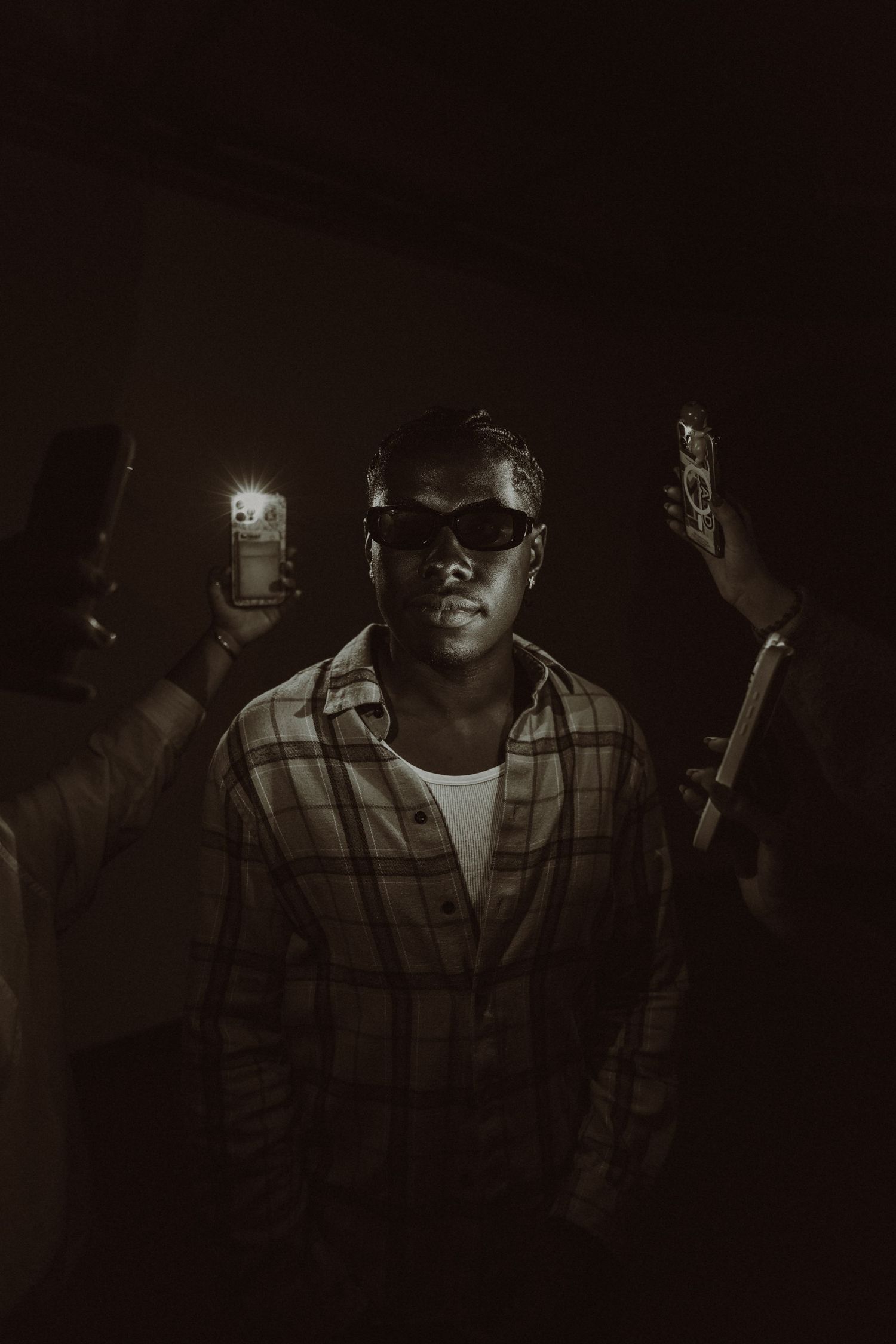 Monochrome portrait of a young man in sunglasses and plaid shirt, surrounded by hands holding glowing phones in dark.