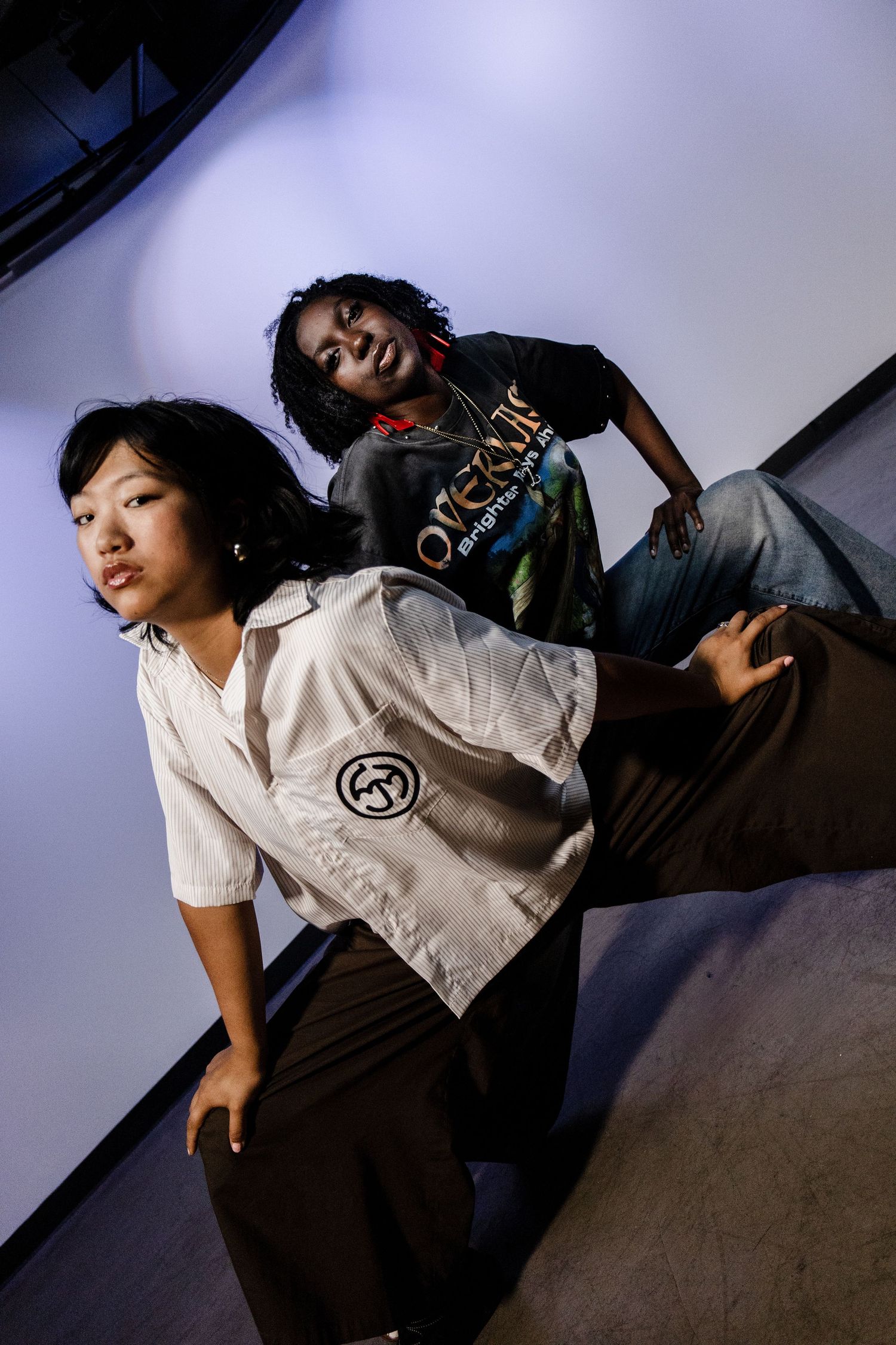 Two young women pose dramatically in streetwear fashion against a purple-lit studio backdrop.