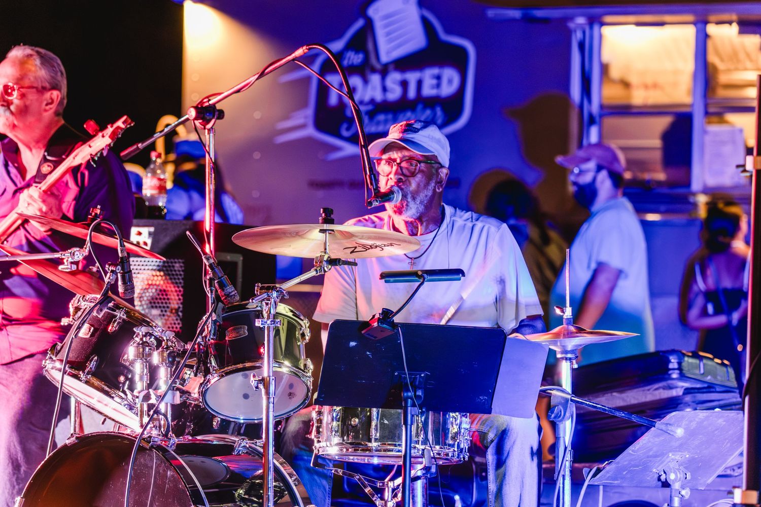 Live band performing at night near a food truck, featuring drummer and guitarist on stage.