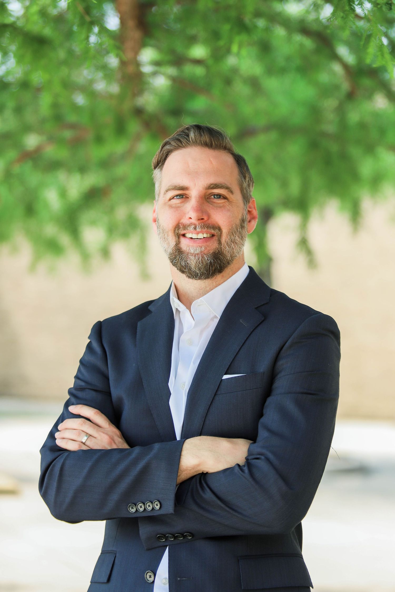 Smiling bearded man in navy suit stands with arms crossed outdoors under green tree foliage.