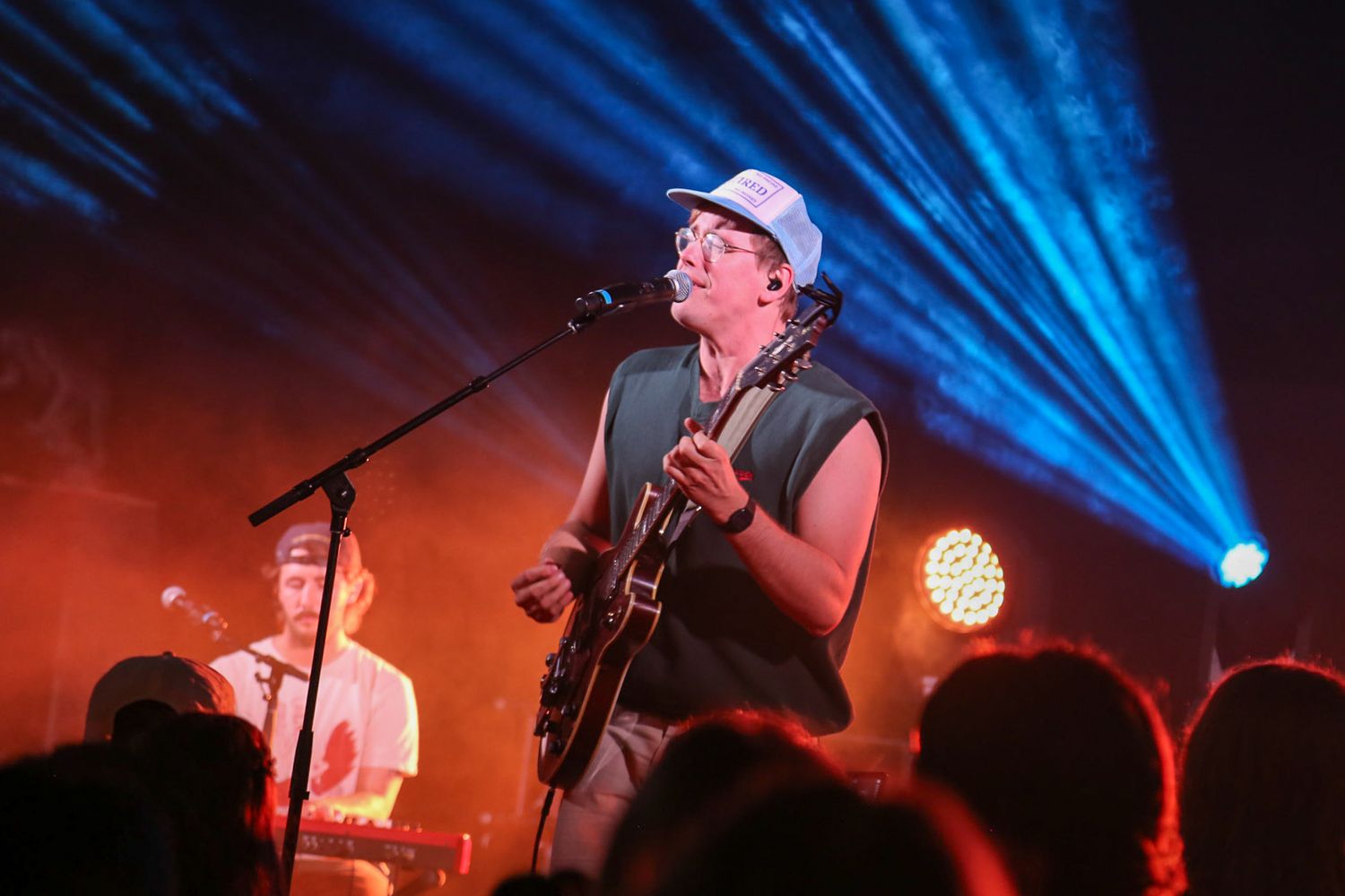 Guitarist in white cap performs on stage with dramatic blue and red lighting at a live music concert.