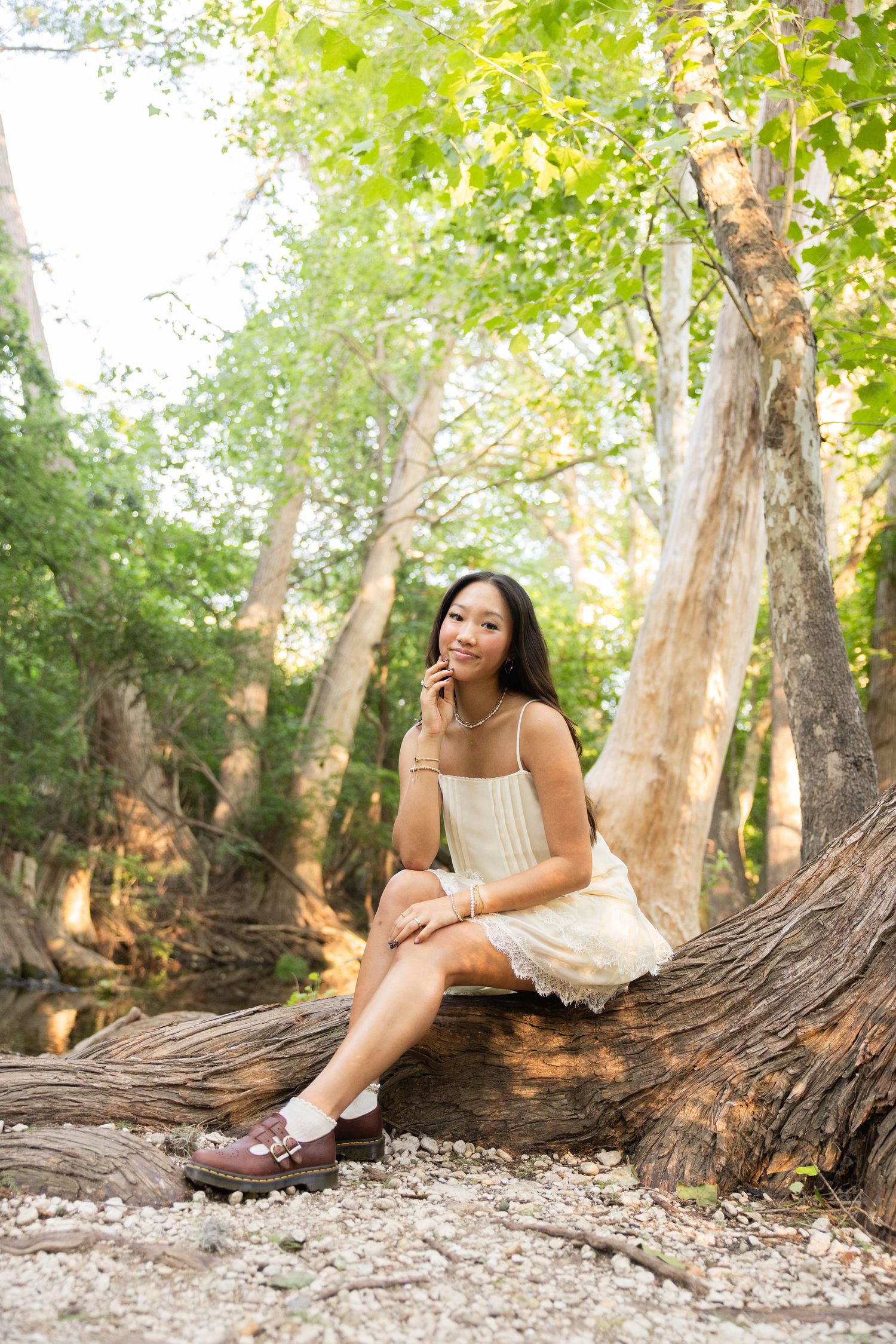 Young woman in white dress sits on fallen tree trunk in a sunlit forest.