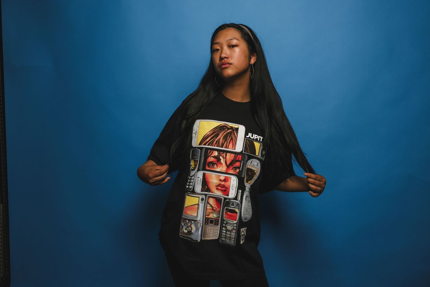 Young woman in oversized black JUPITTXR graphic t-shirt poses against vibrant blue background.