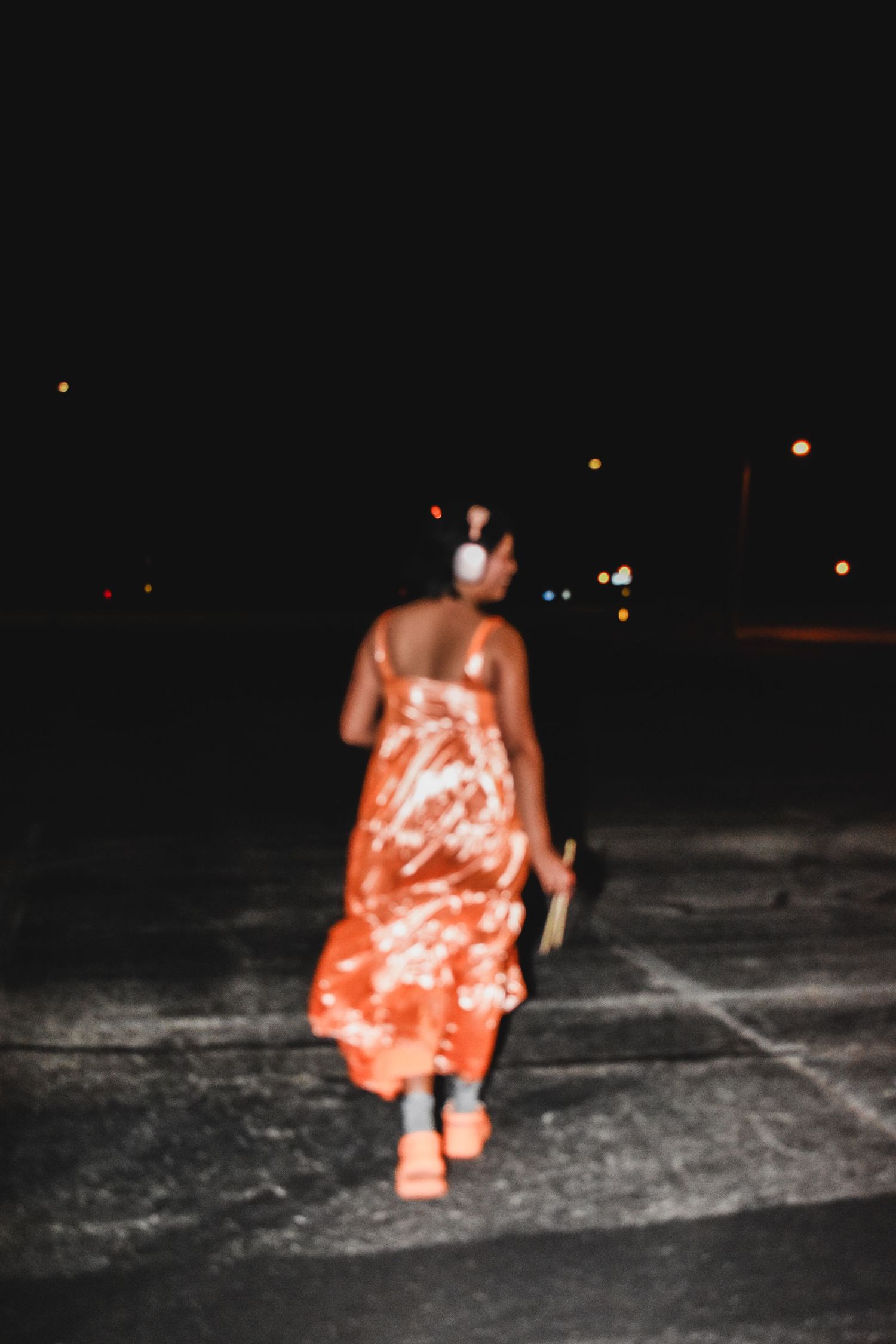 A woman in an orange floral dress and white headphones walks away through a dark parking lot at night.
