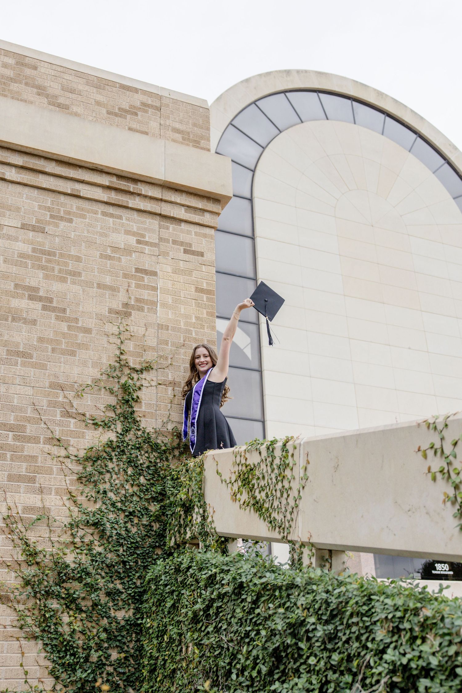 Graduate in cap and gown raises mortarboard triumphantly atop a university building surrounded by ivy.