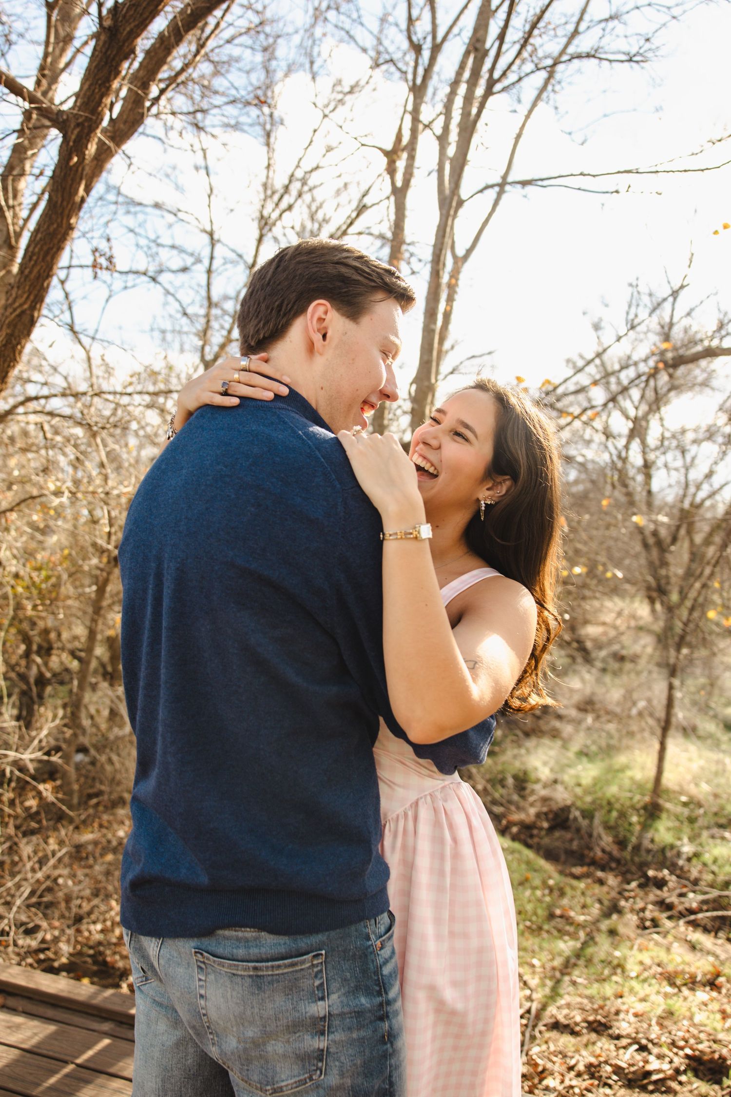 Happy couple laughing and embracing outdoors among bare trees in golden sunlight.