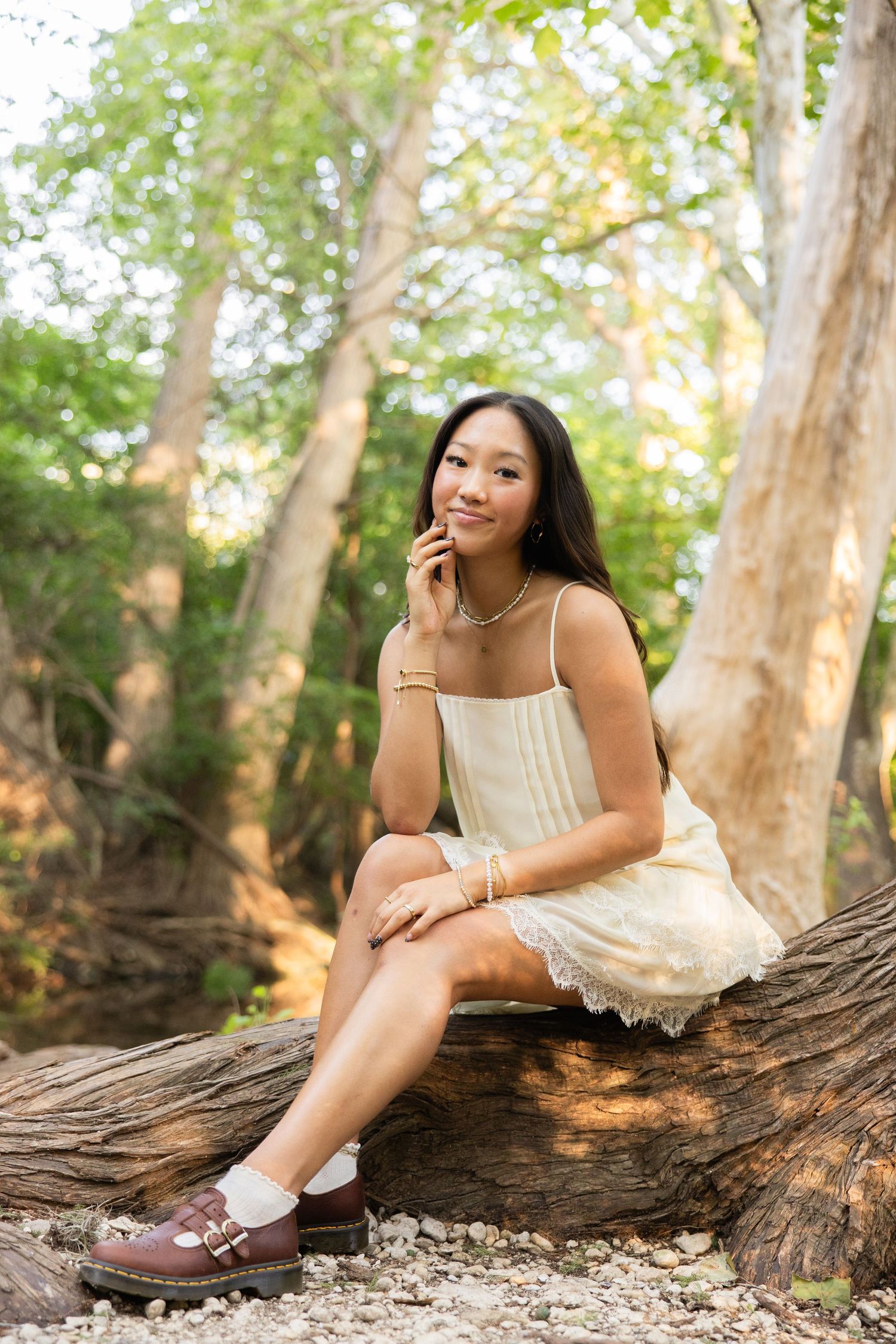 Young Asian woman in cream dress sitting on log in lush green forest setting.