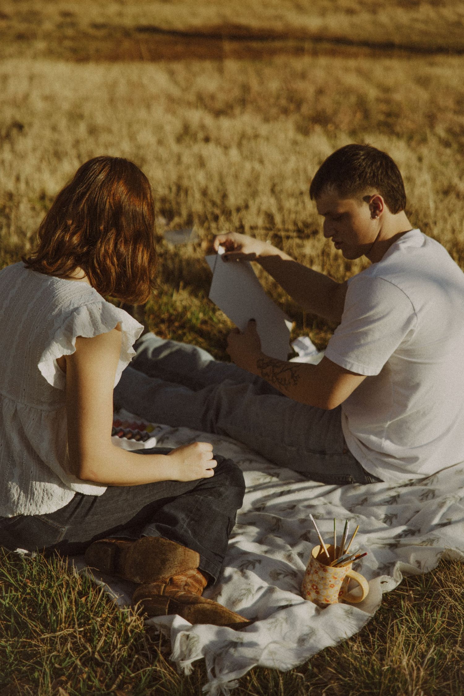 Couple sitting on a blanket in a golden field, sketching and painting together at sunset.