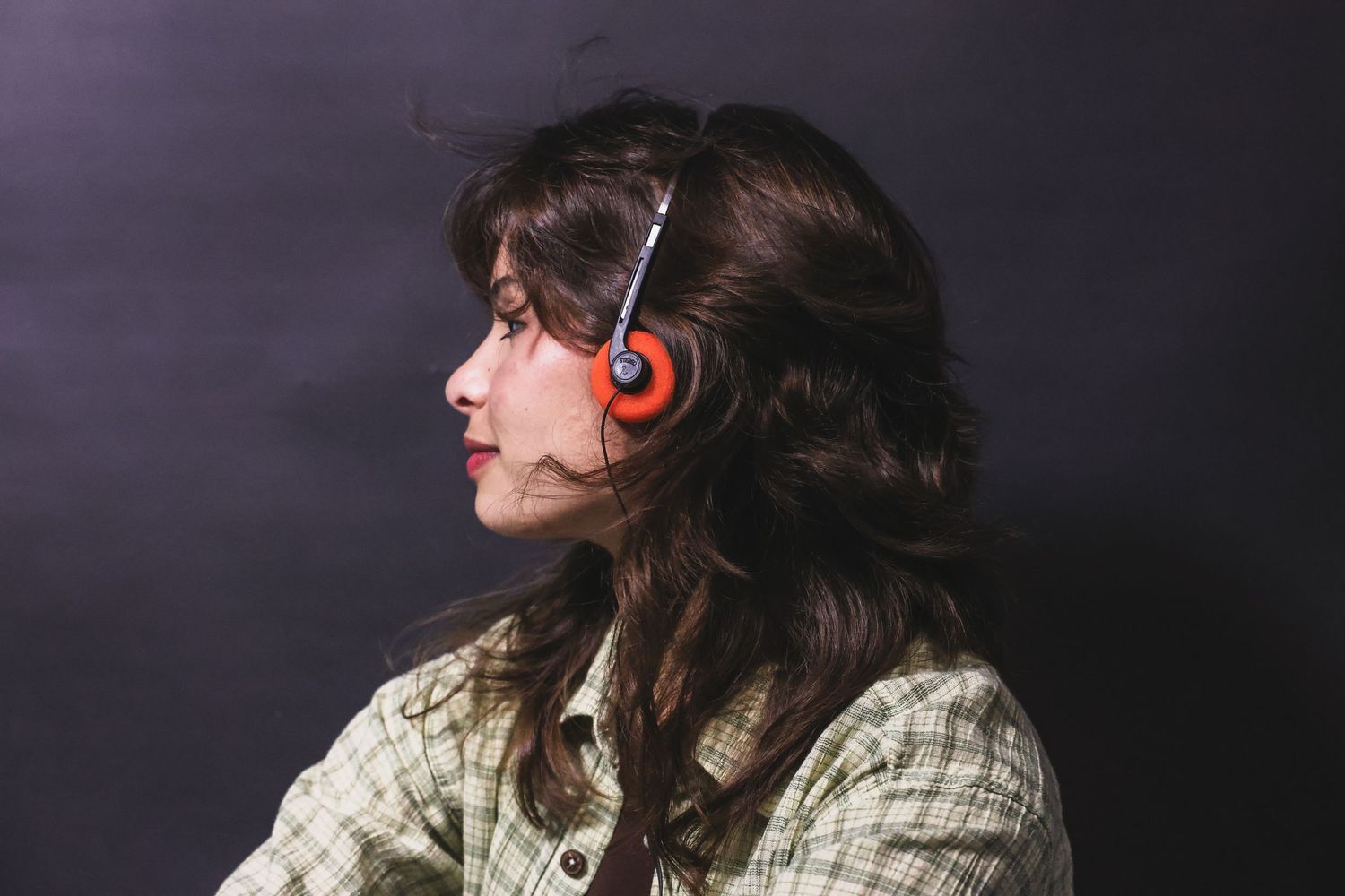 Young woman with dark hair wearing orange headphones and plaid shirt against dark background.