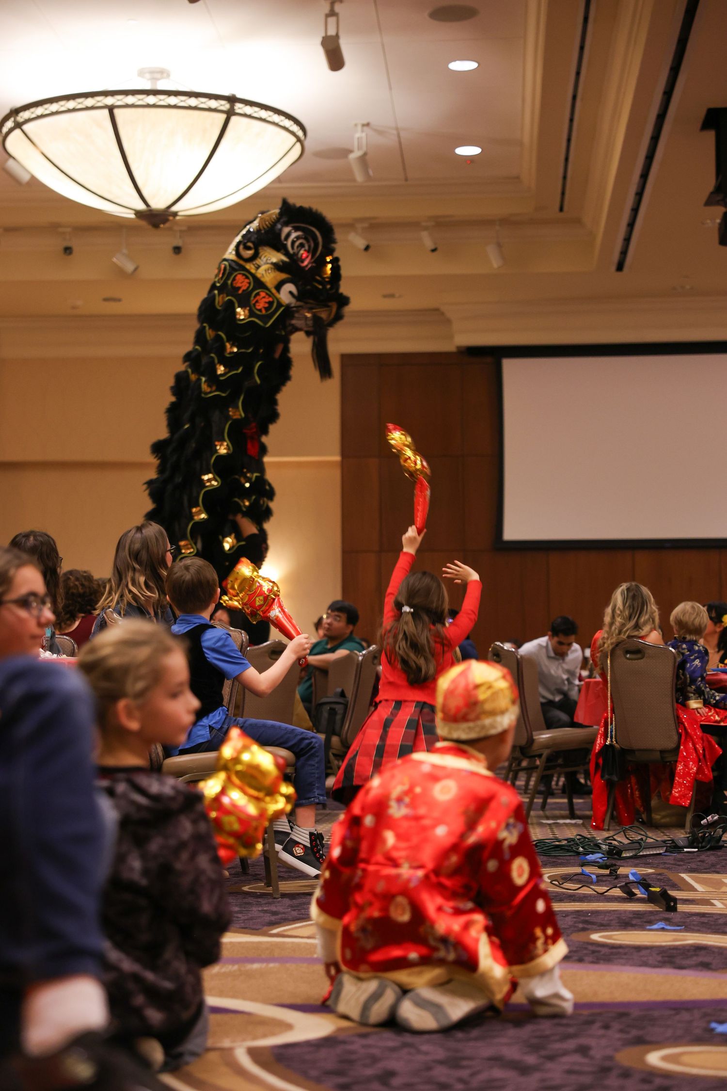 Children in colorful costumes interact with a black Chinese lion dancer during a festive cultural performance.