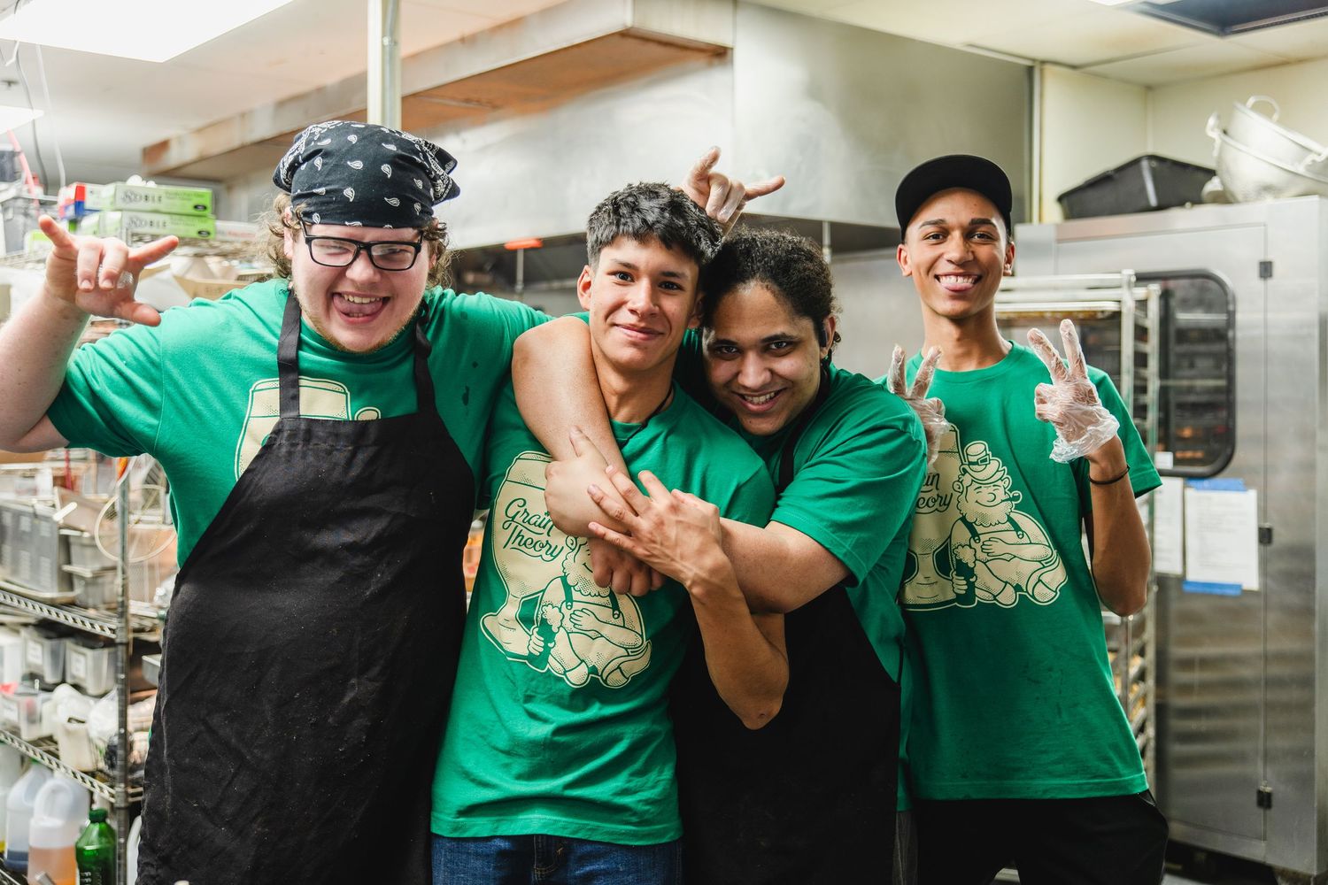 Four smiling kitchen staff in green t-shirts pose together in a restaurant kitchen.